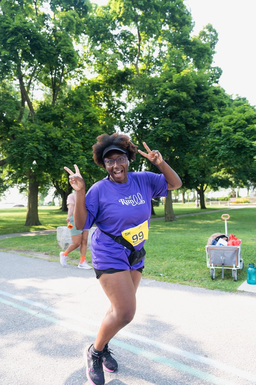 A woman running in a 5K race, smiling and making peace signs with both hands, wearing a purple race T-shirt with the text 'Little Ranged' and race bib number 99, in a park with green trees in the background.
