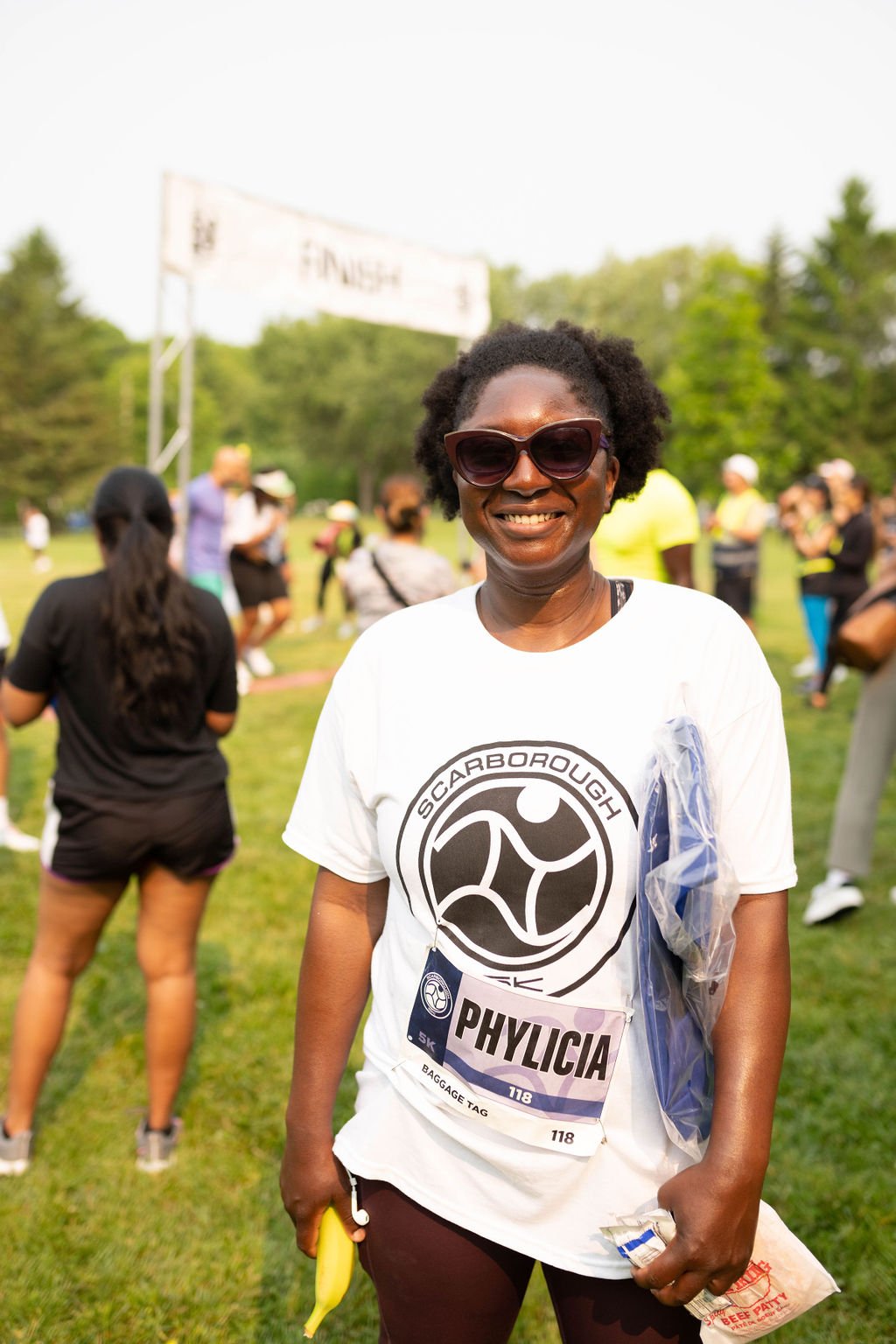 Smiling woman wearing sunglasses and a Race bib with the name 'PHYLICIA' at an outdoor event, with other people in the background near a finish line on a grassy field.