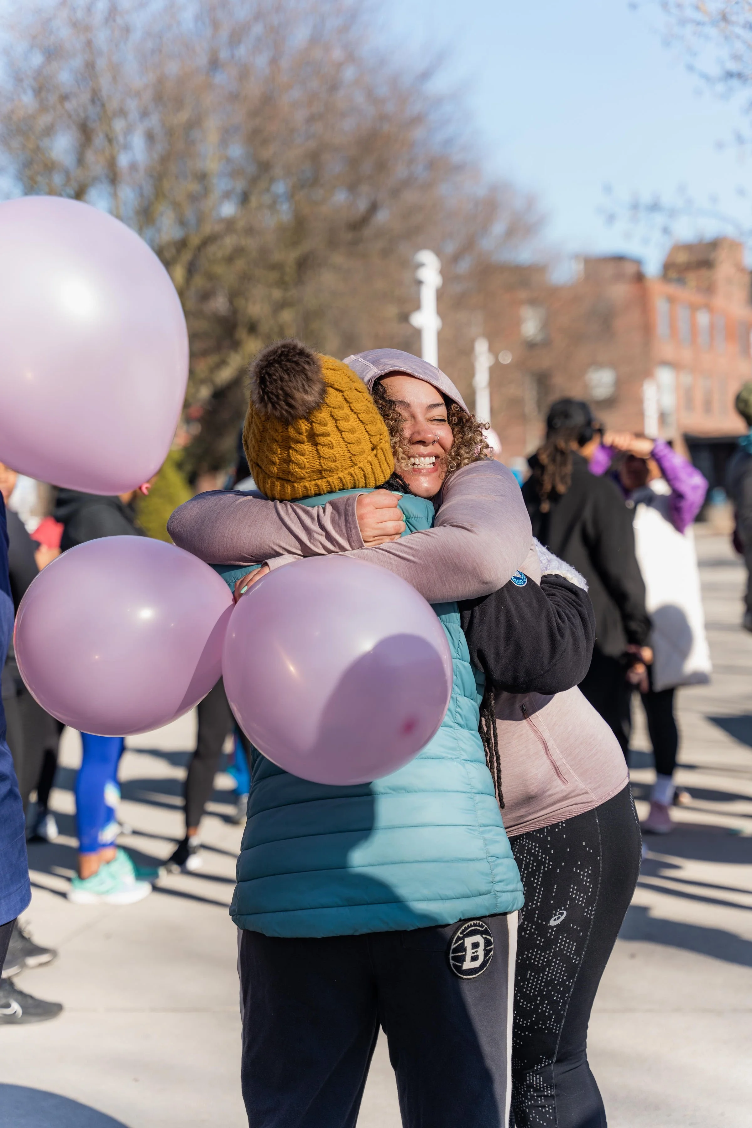 Two women hugging during an outdoor event, one wearing a yellow knit hat with a pom-pom and holding pink balloons.