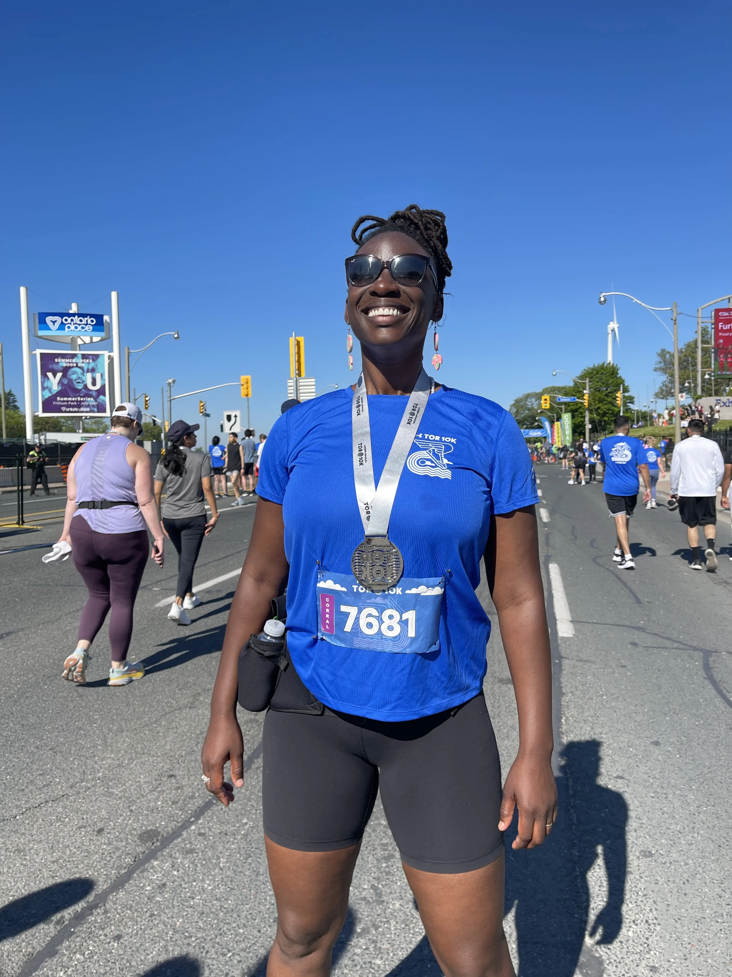 A woman in a blue race shirt with a medal around her neck, standing on a city street after a race, smiling with sunglasses, with other race participants and spectators in the background on a sunny day.