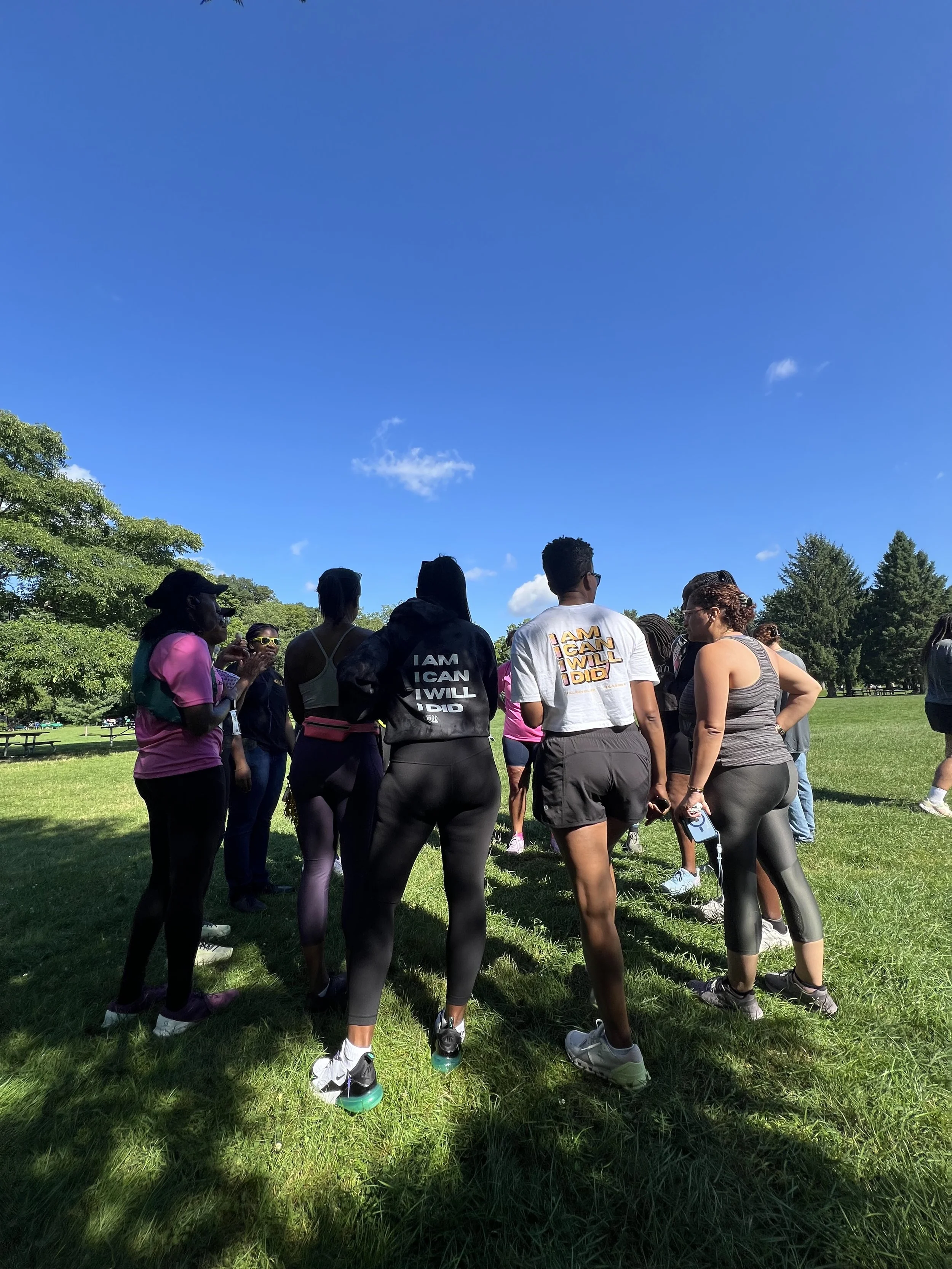 Group of people gathered outdoors on a grassy field on a sunny day with clear blue sky and trees in the background.