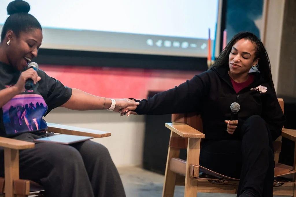 Two women sitting on wooden chairs, holding hands, one with a microphone, in a casual indoor setting.