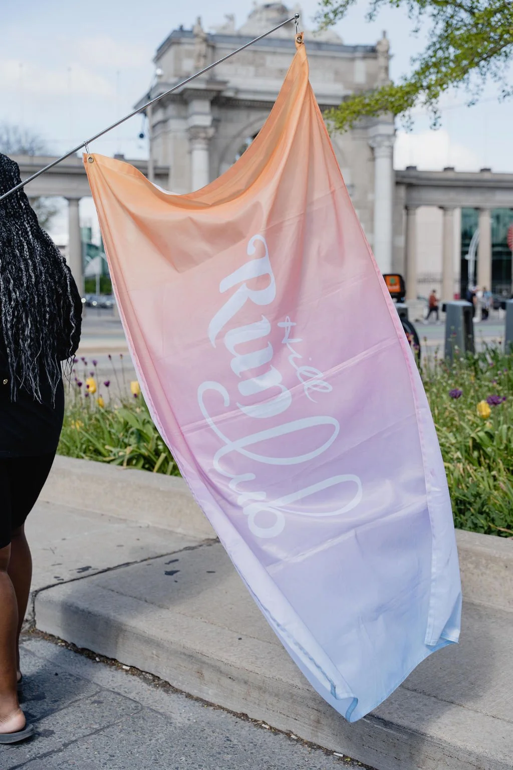 A large, pastel-colored flag with the words 'xoxo true Love' written on it, held by a person on a city sidewalk, with a large stone building and flowers in the background.