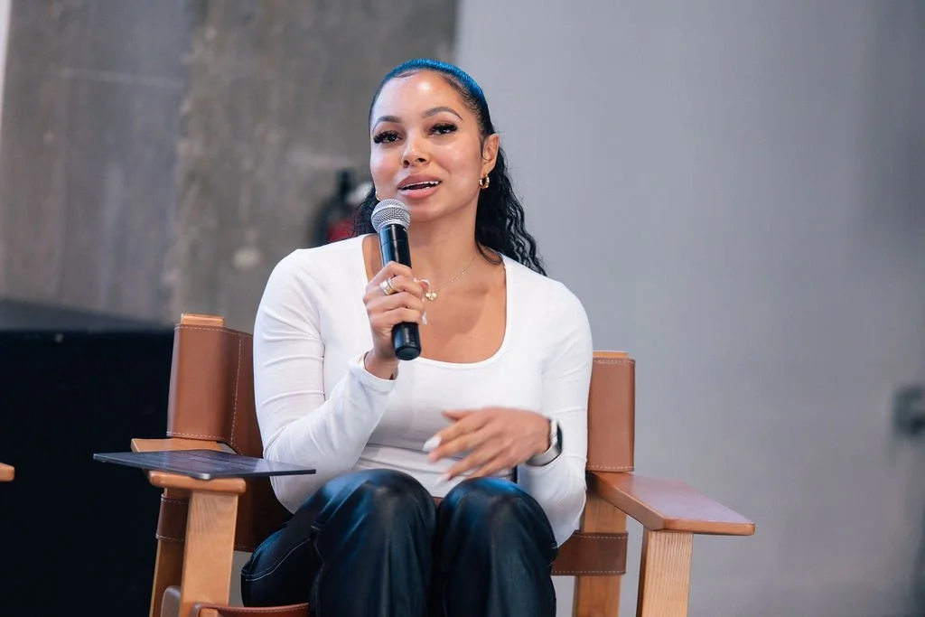 Woman sitting on a wooden chair, holding a microphone, speaking during a panel discussion or interview, with gray wall background.