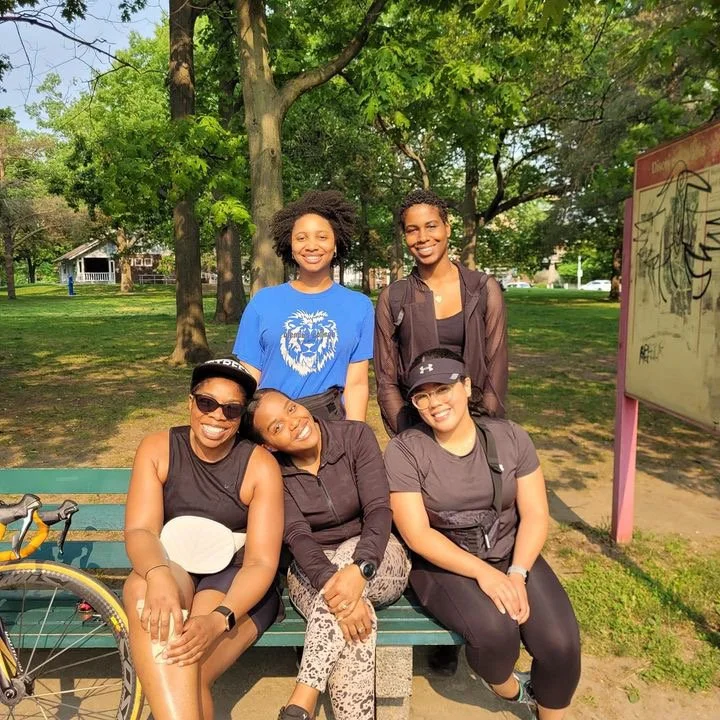 Five women sitting and standing on a park bench, smiling. They are outdoors with trees and a grassy area in the background.