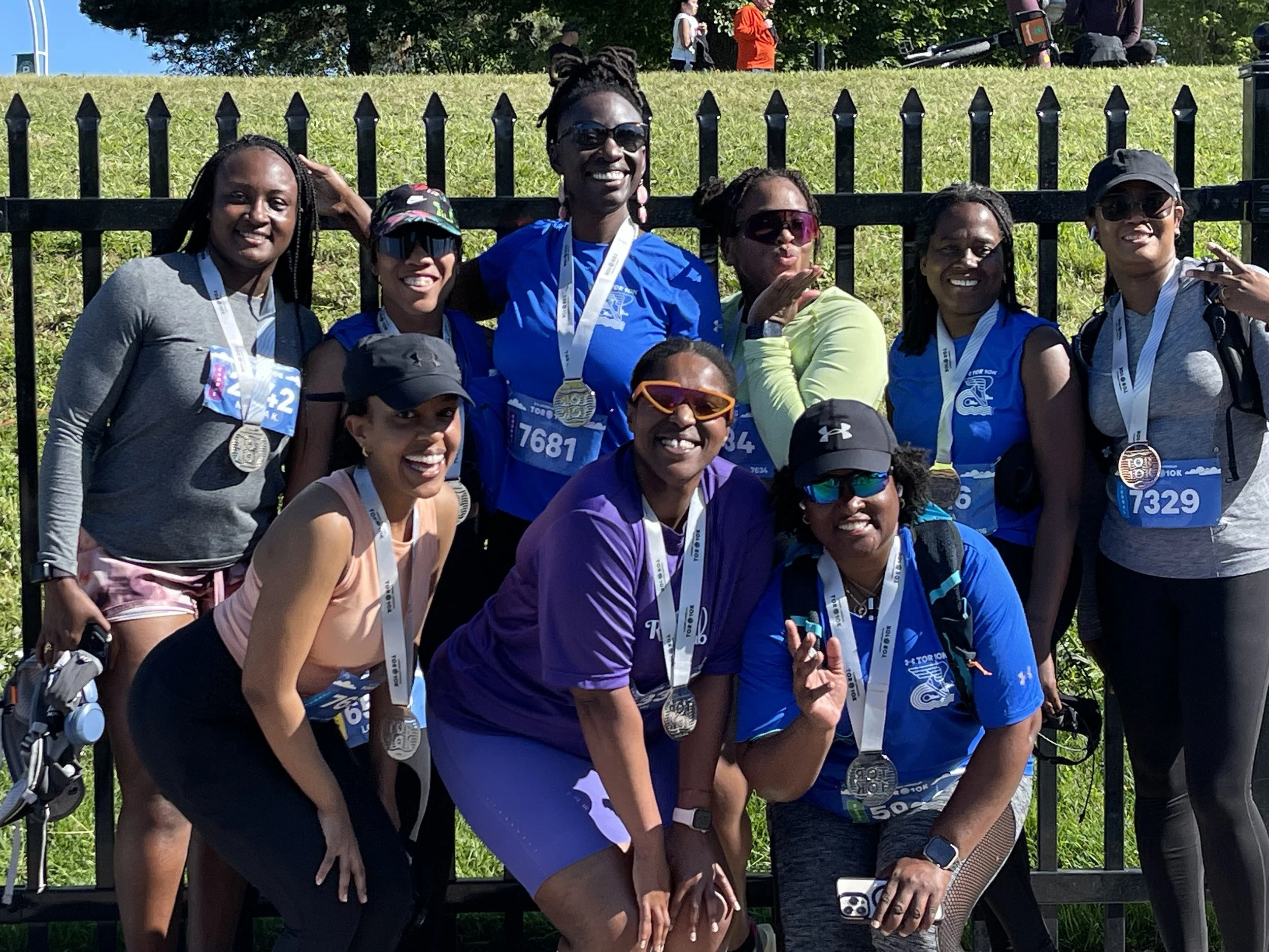 A group of eleven women at a running event wearing medals, athletic gear, and race bibs, standing outdoors in front of a black fence, smiling and posing for the photo.