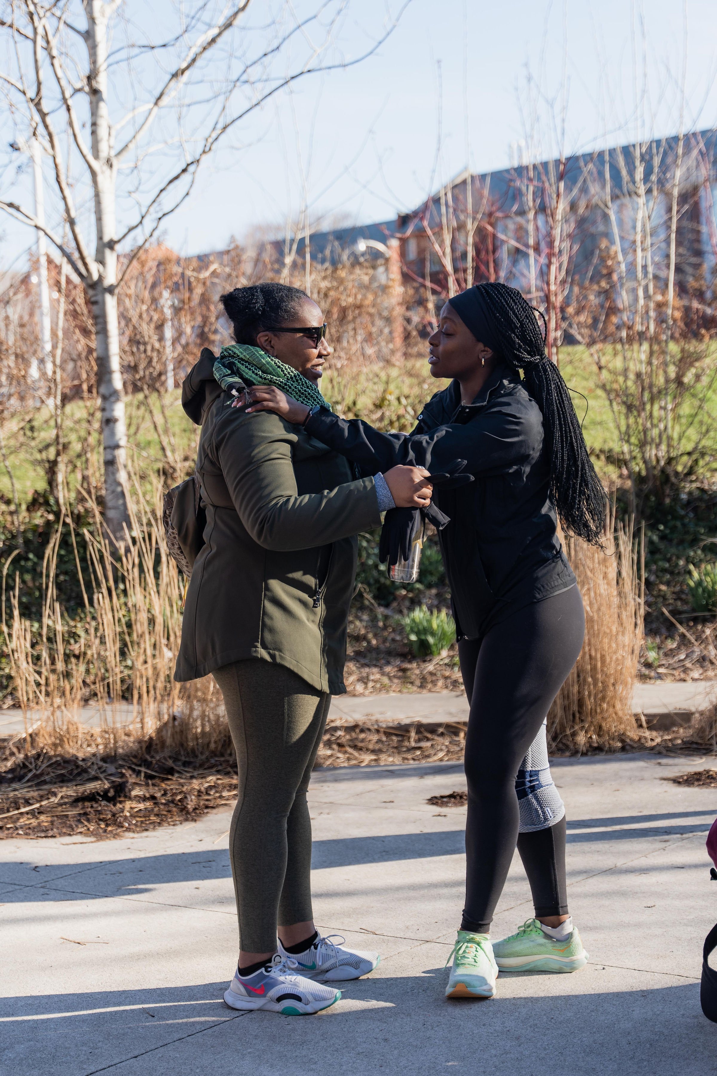 Two women interacting outdoors on a sunny day, one wearing sunglasses and a green scarf, the other with braided hair and black athletic clothing, standing on a sidewalk with dry plants and leafless trees in the background.