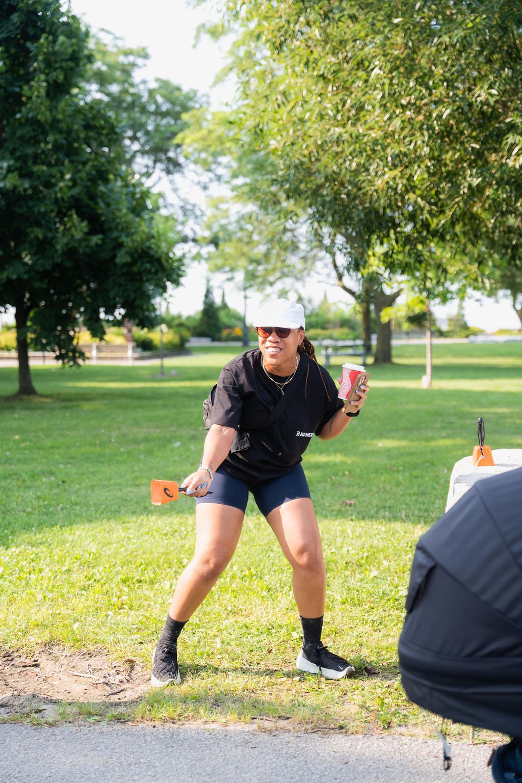 Woman wearing black athletic outfit, white cap, and sunglasses, holding a disposable coffee cup and a small orange shoveling tool, smiling and posing outdoors on grassy area with trees in background.