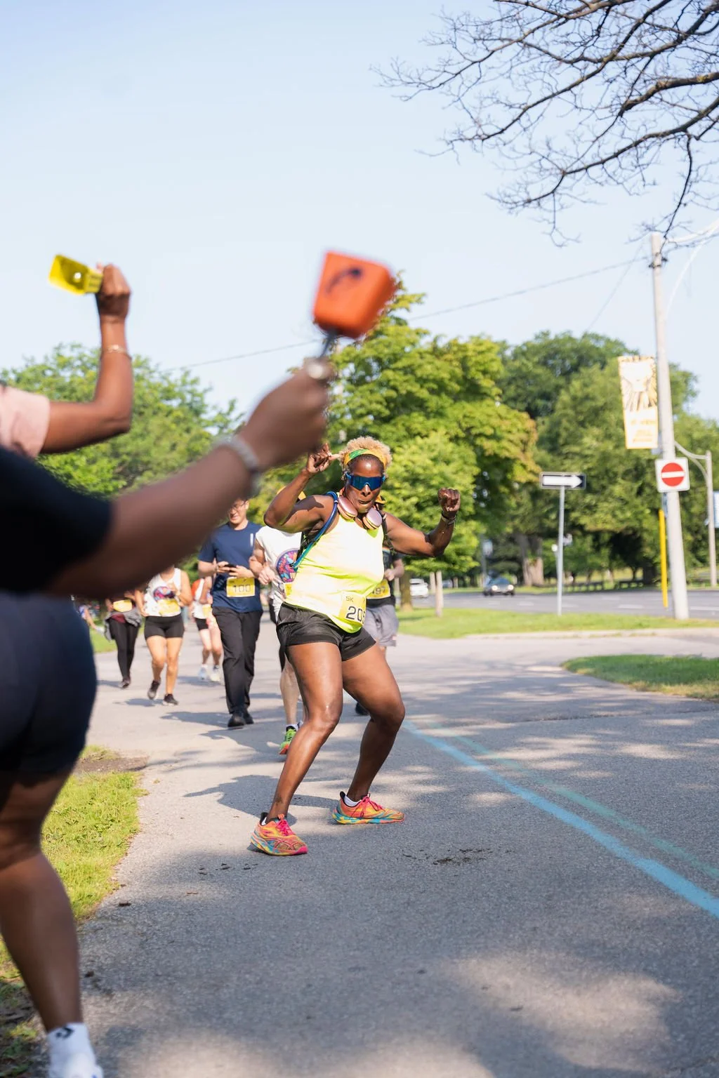 A woman wearing sunglasses, a yellow tank top, and black shorts is running and celebrating during a race, with other runners behind her on a sunny day.