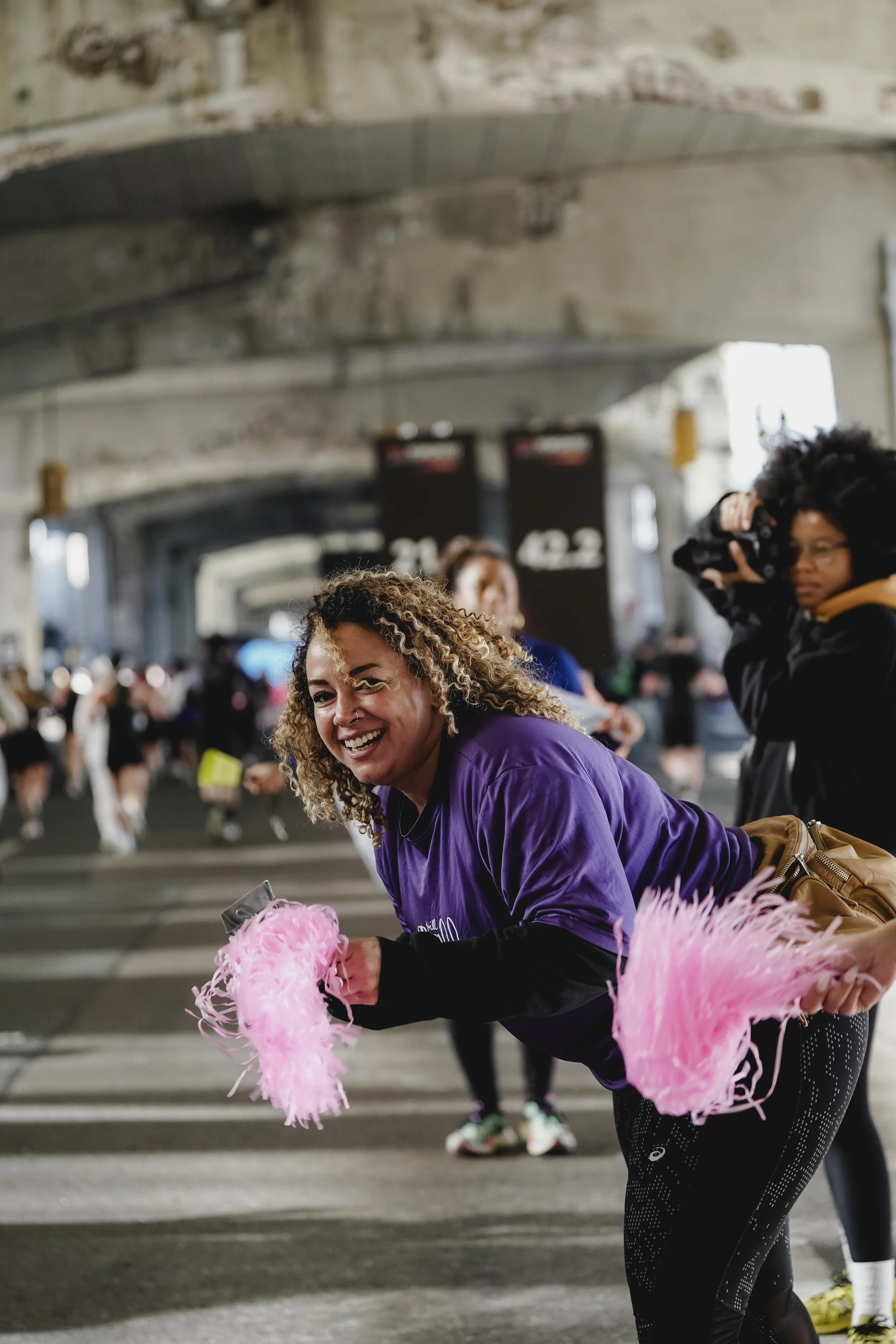 A woman smiling and holding pink feathered pom-poms at a crowded indoor event under a bridge or tunnel.