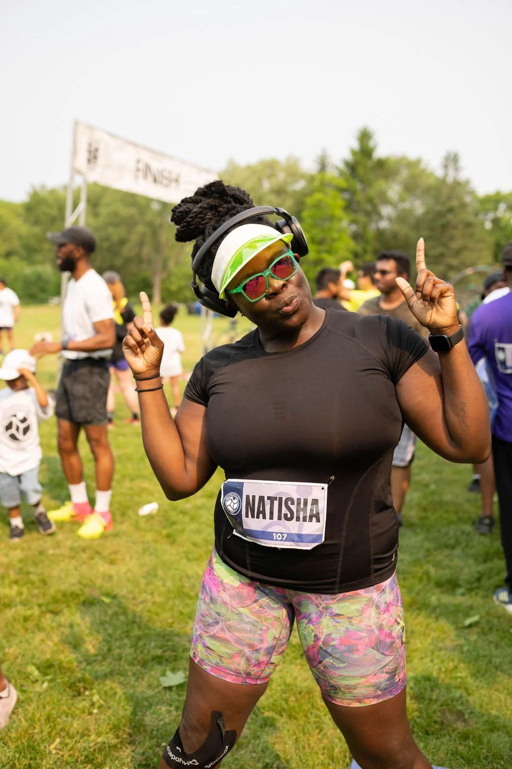 A woman wearing sunglasses, headphones, and sports gear at a race event, standing on grass with other participants and a finish line in the background.