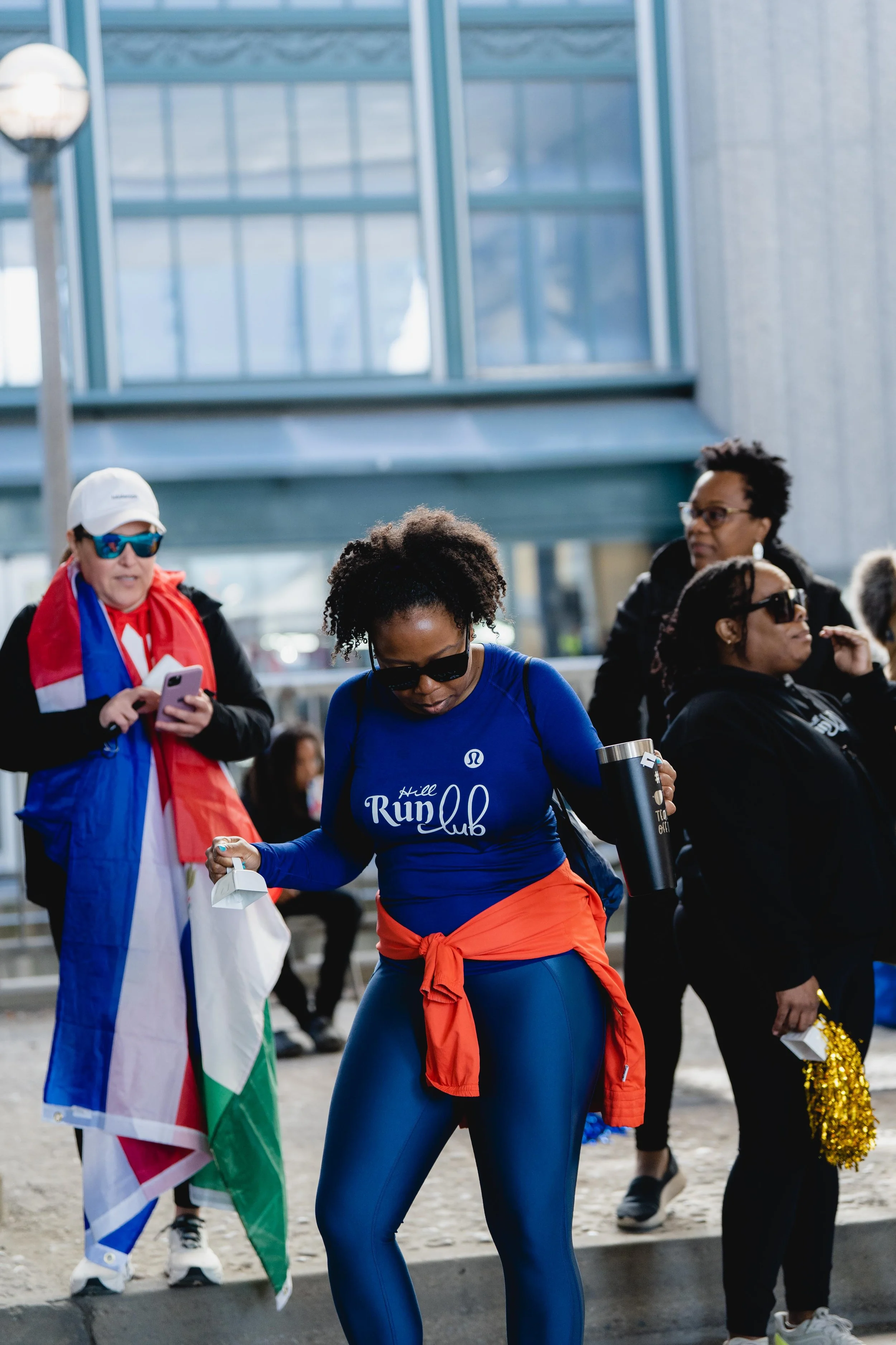 A group of people gathered outdoors near a modern glass building, with a woman in athletic wear, a blue long-sleeve shirt, and sunglasses in the foreground, holding a water bottle.