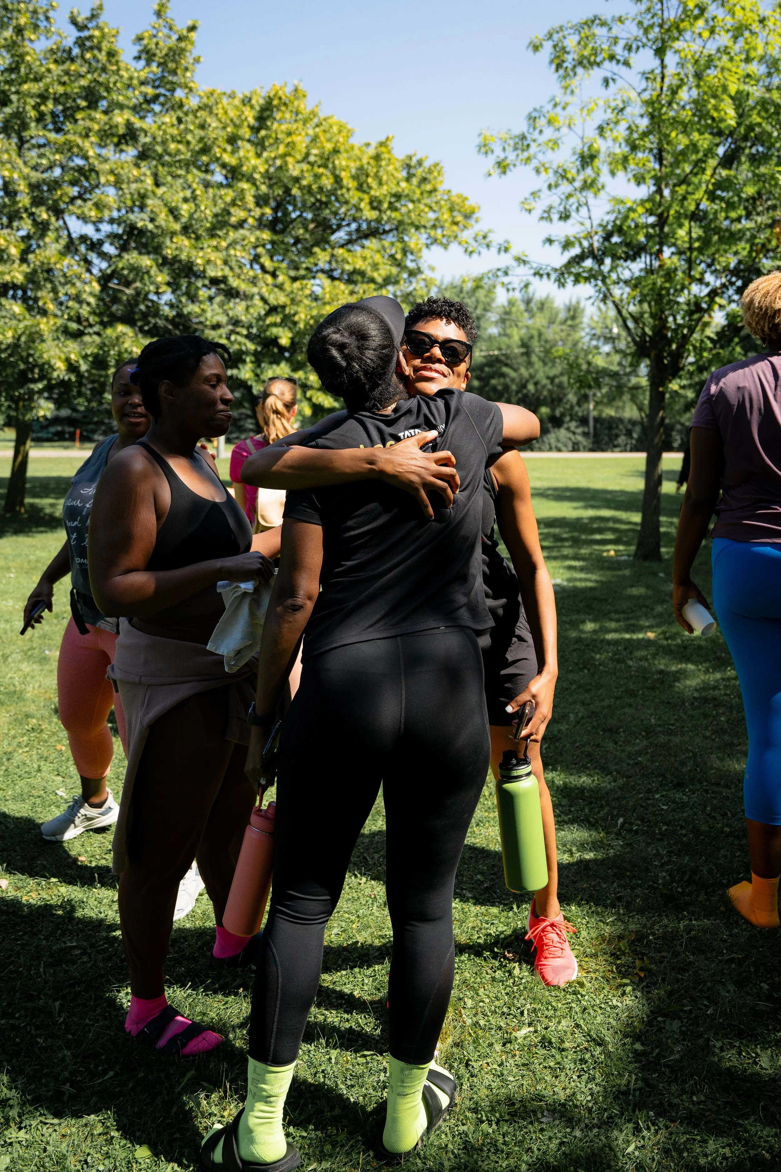 Two women hugging outdoors in a park with other women nearby, trees, and a grassy field.
