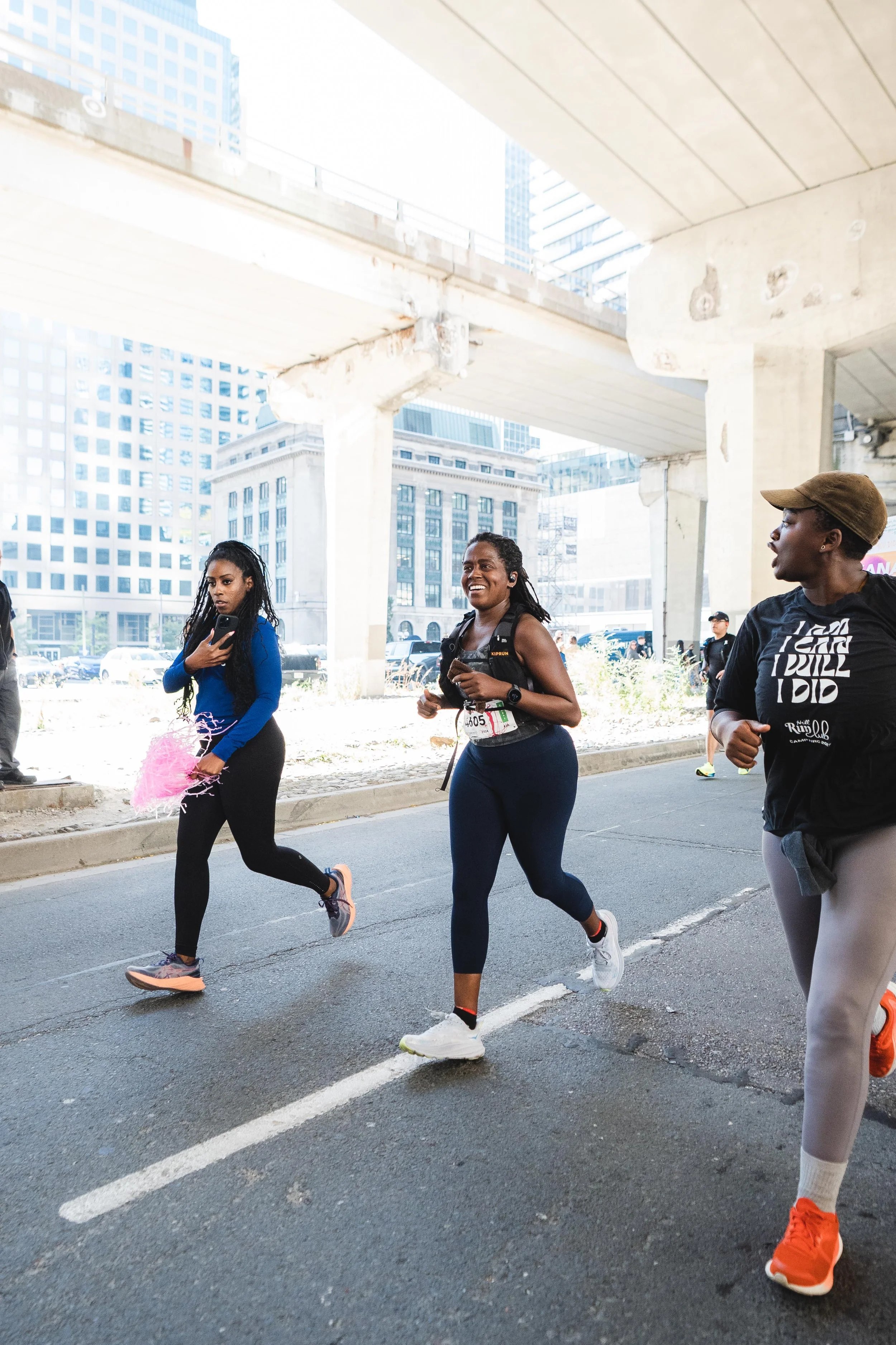 Three women run in a street marathon under an urban overpass, with tall buildings in the background.