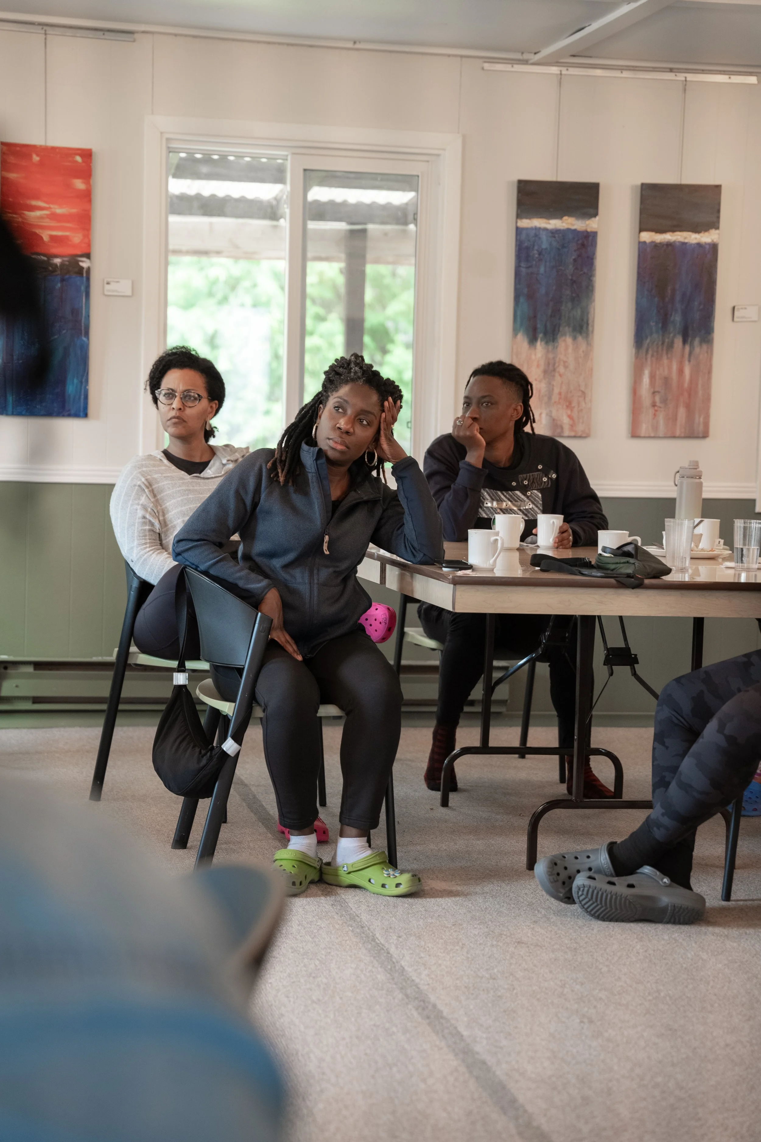 Three women sitting at a table in an art gallery, listening attentively. One woman in the foreground has her hand on her head and wears a dark jacket. The other two women are seated behind her, with one wearing glasses and a striped sweater, and the 