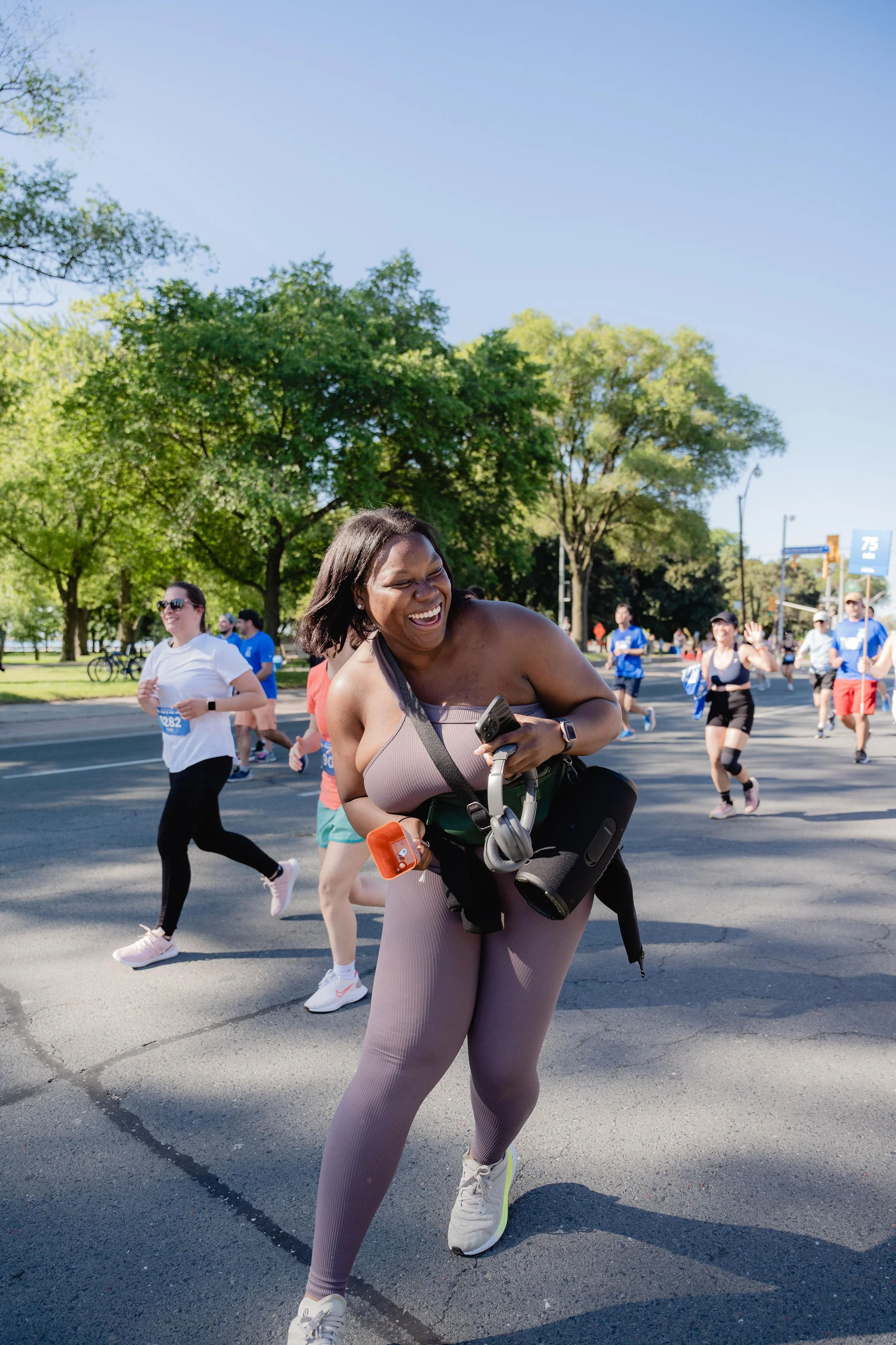 A group of runners participating in a race or marathon, with a woman in the foreground laughing and smiling as she runs on a sunny day with green trees in the background.