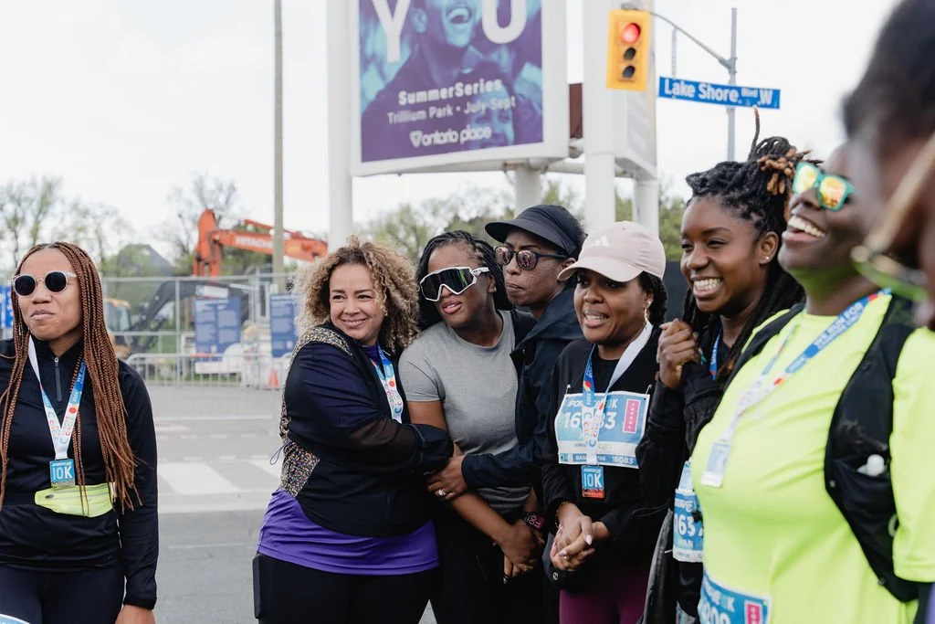 Group of women smiling and standing together at a race event, some wearing bibs and sunglasses, with construction and a billboard in the background.