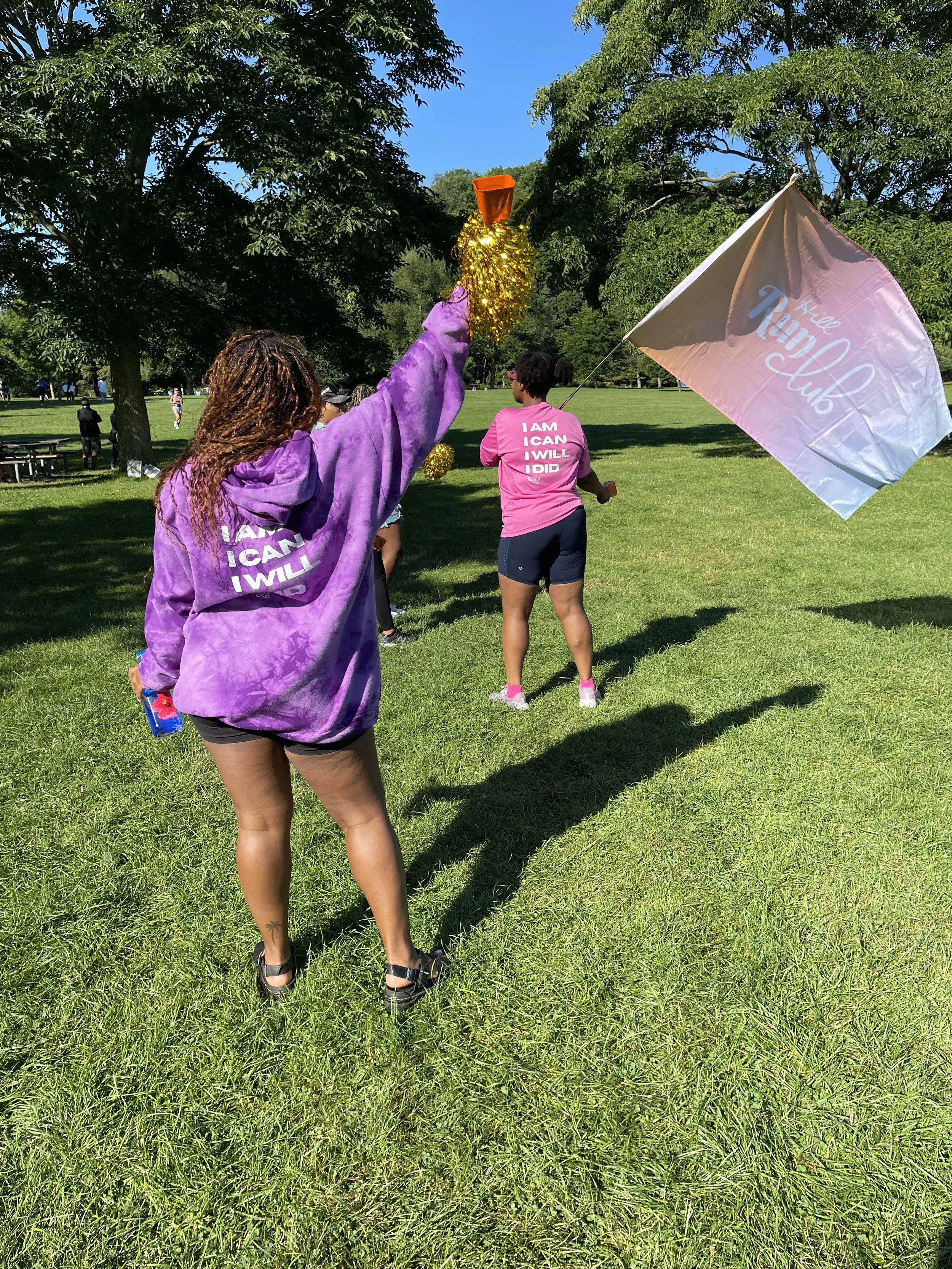 People participating in an outdoor event in a park, some holding a flag that says 'Hera Club' and wearing pink shirts with motivational text.