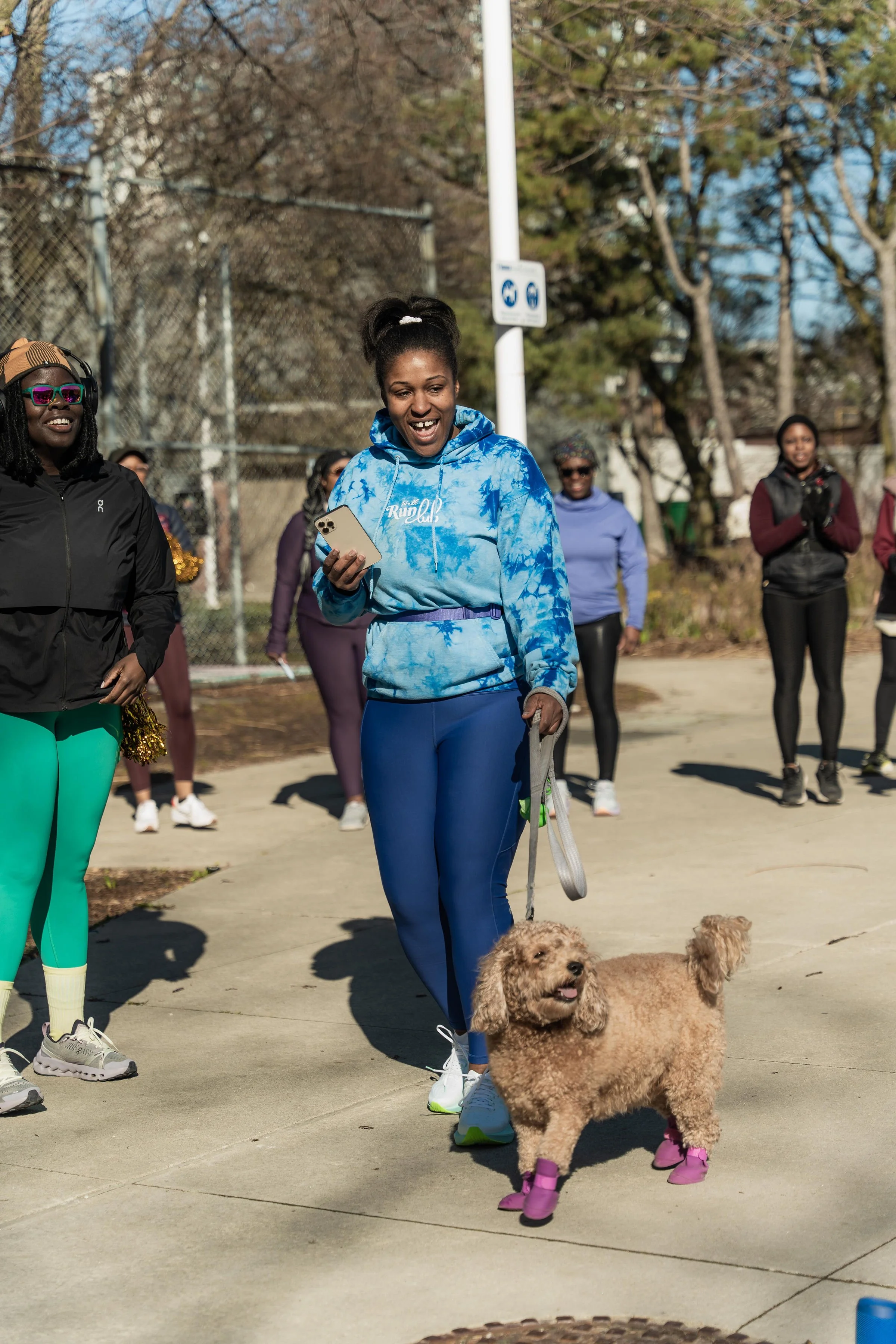 Smiling woman in blue tie-dye hoodie walking her dog with pink booties at outdoor event, surrounded by other women.