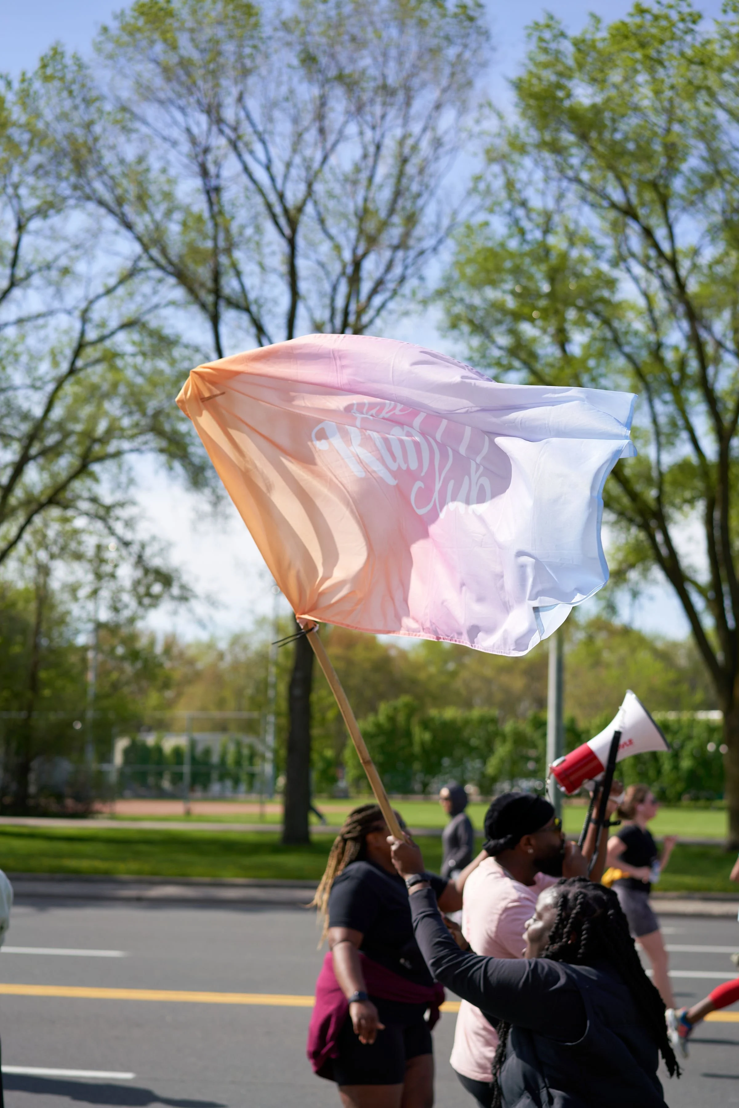 People participating in a march or rally, some holding flags and banners, on a street with trees in the background.