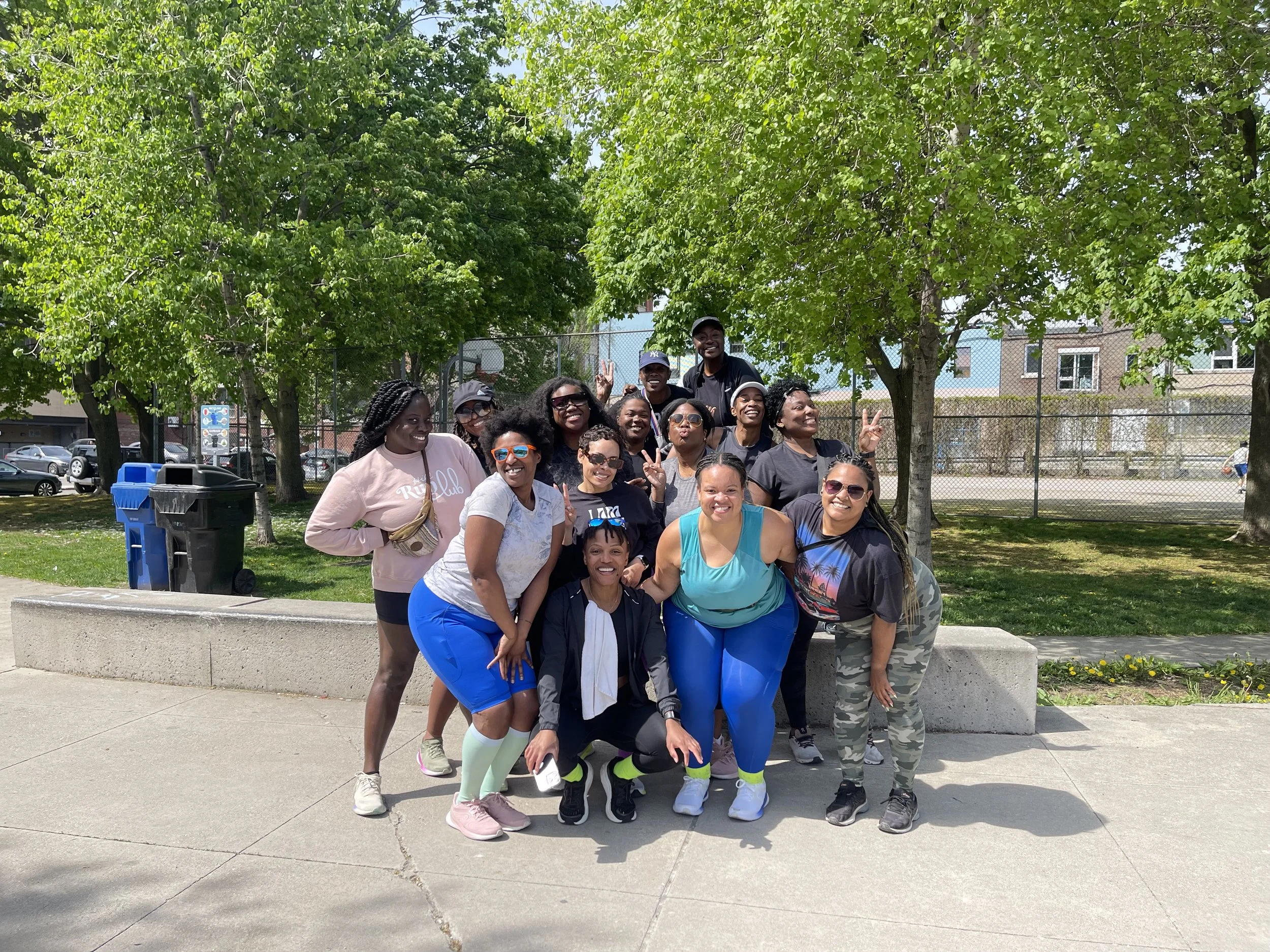 Group of people outdoors posing for a photo under green trees, some showing peace signs and smiling.