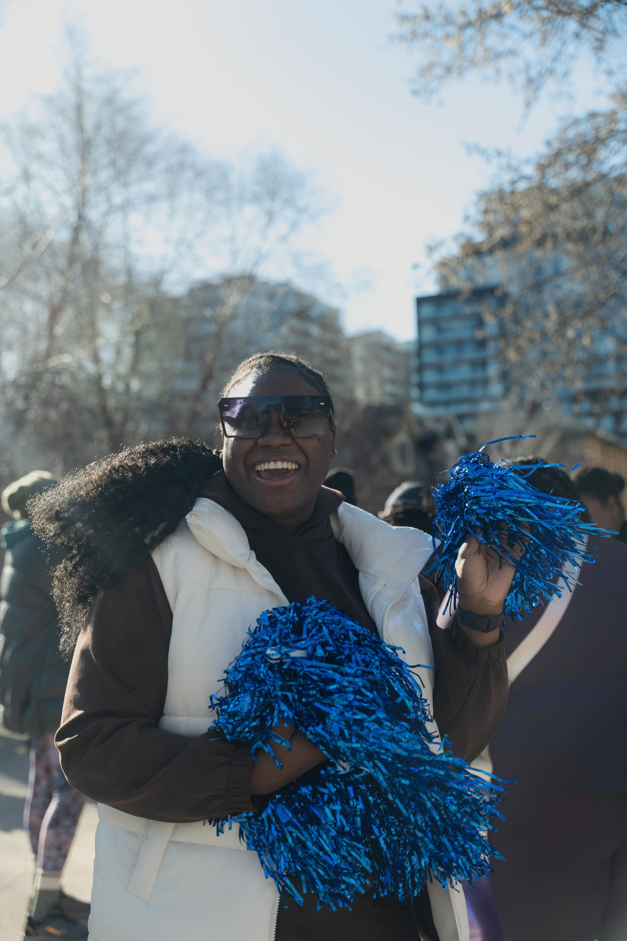 A woman outdoors, smiling and holding blue pom-poms, wearing sunglasses and a white vest, during daytime with buildings and trees in the background.