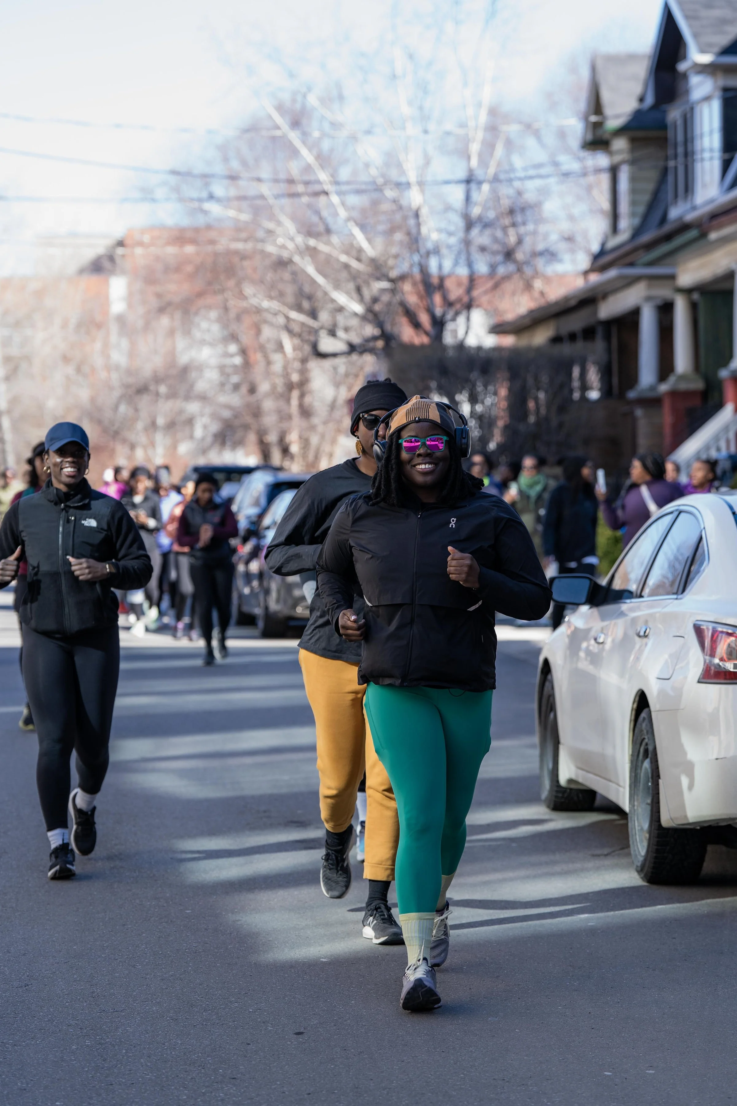 A group of people jogging on a city street during daytime, with houses and parked cars in the background.