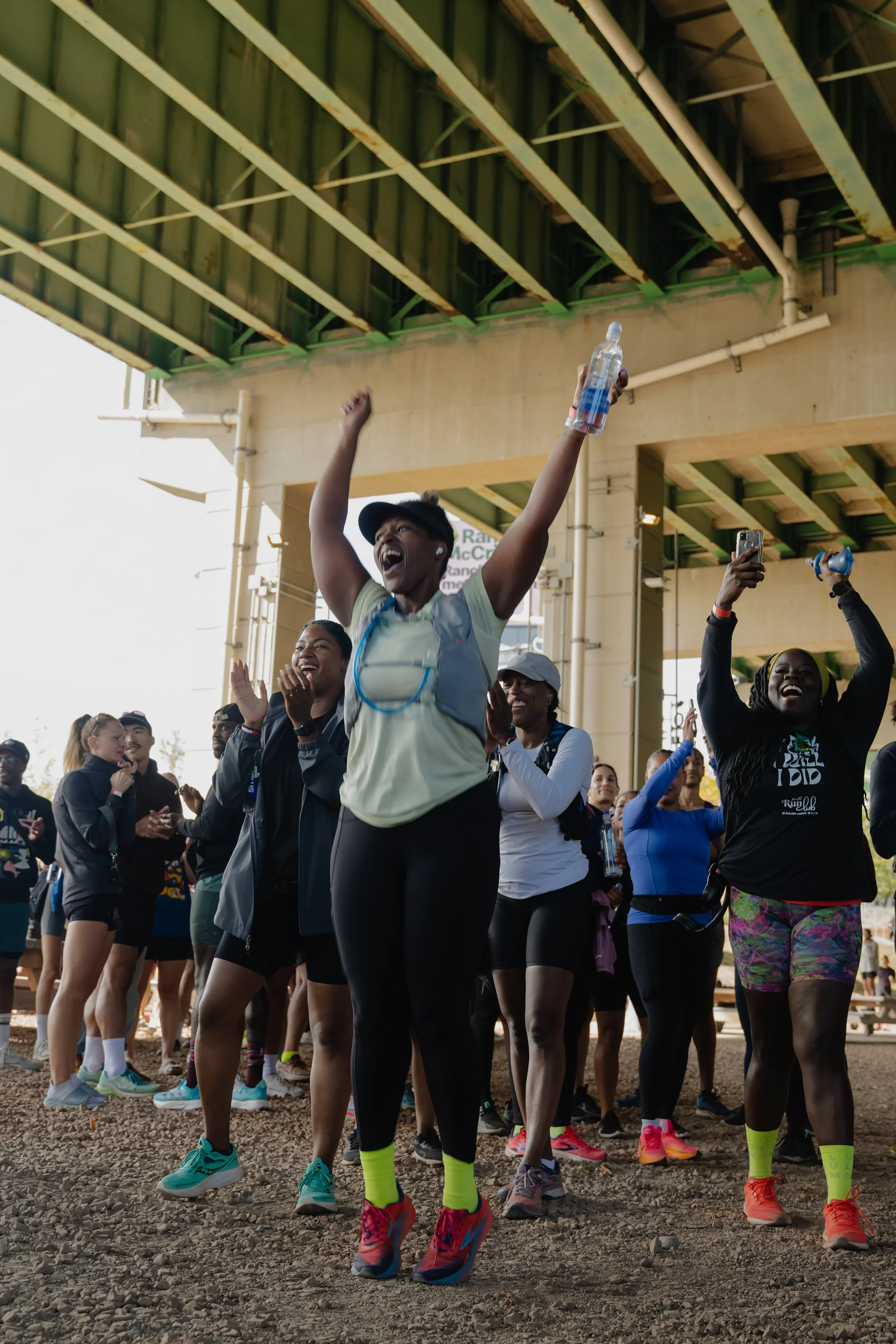 Group of runners celebrating after finishing a race underneath a bridge, with some raising their hands and smiling.