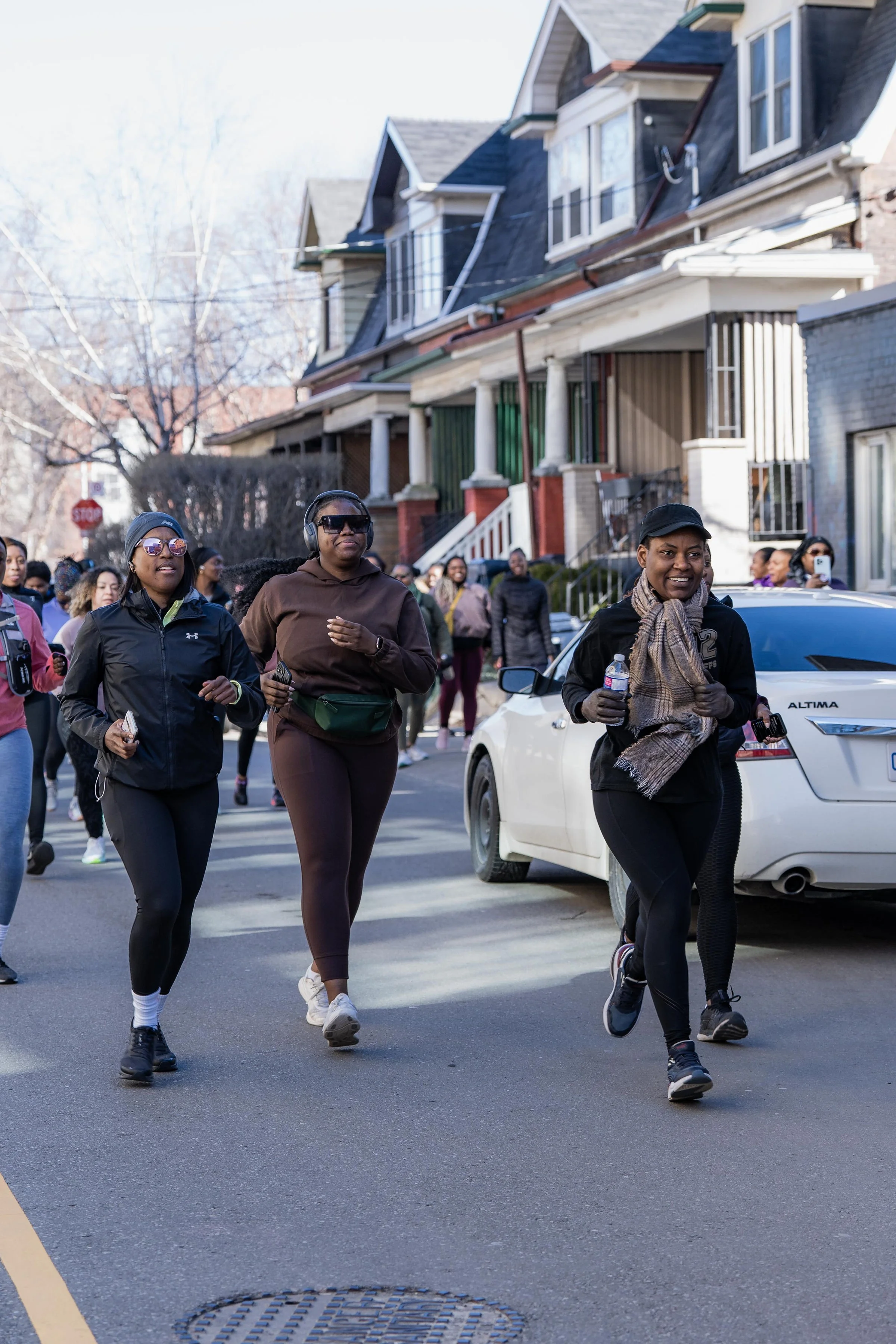 A group of people, mostly women, jogging and walking down a suburban street on a clear day, with houses and trees in the background.