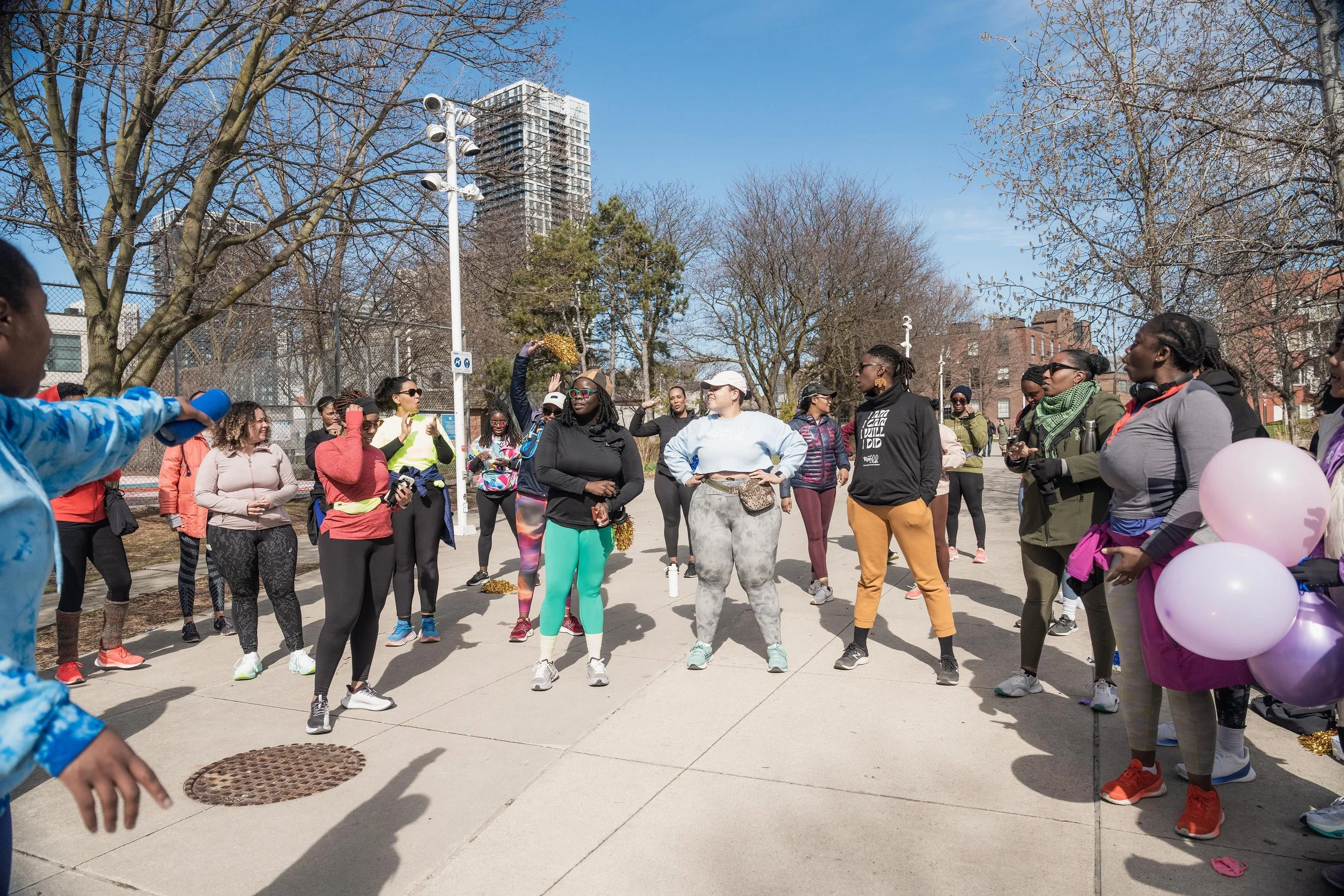 Group of women gathered outdoors on a sunny day, some wearing athletic clothing, participating in a fitness or community event in a city park.