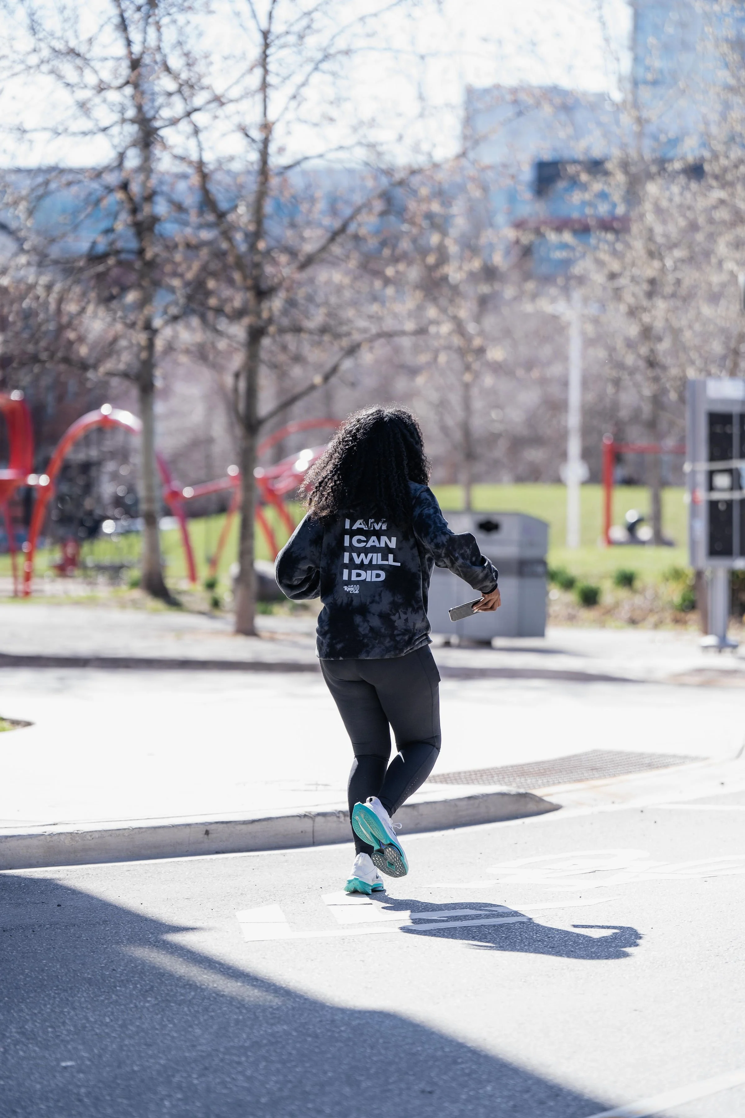 A woman running outdoors on a sunny day, holding a smartphone in her hand, with trees and playground equipment in the background.