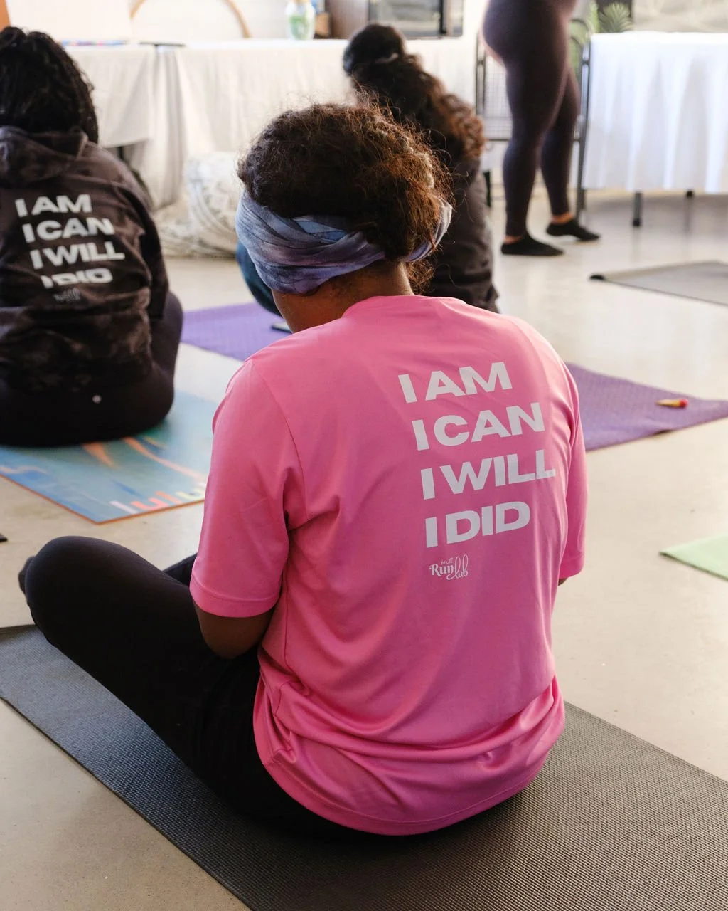 A woman sitting on a yoga mat during a fitness class, wearing a pink shirt with motivational text. She is facing away, and other participants are visible around her, also seated on mats.