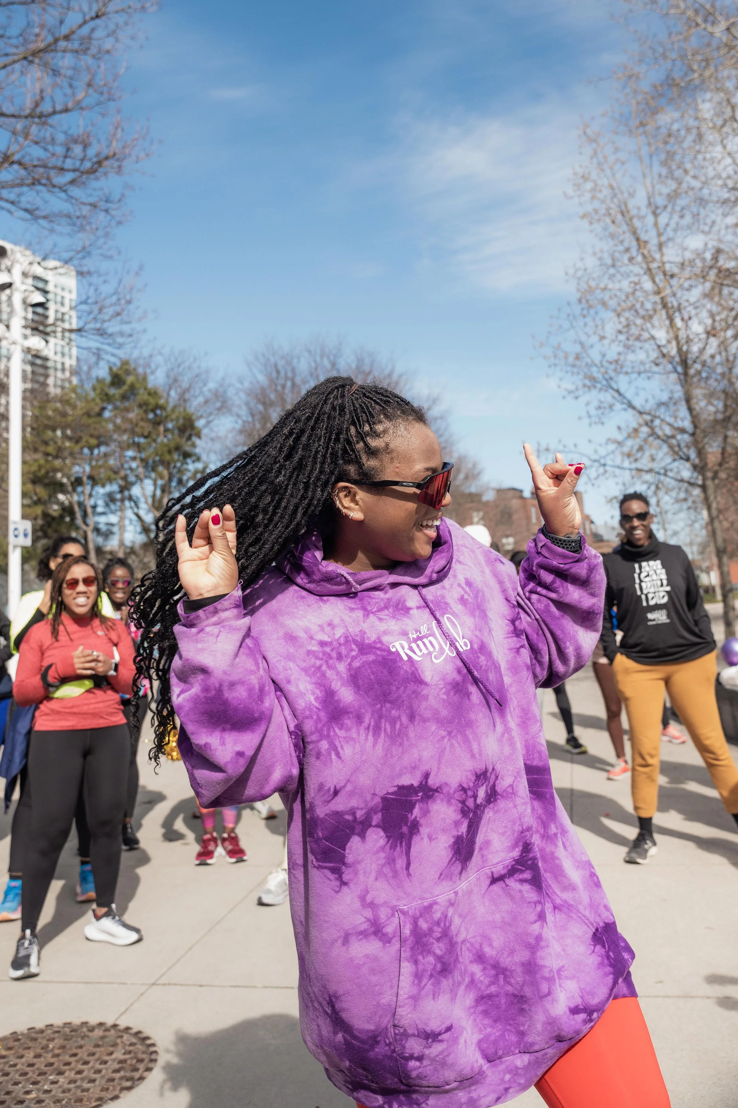 A woman dancing outdoors with a group of people, wearing a purple tie-dye hoodie and sunglasses, on a sunny day with trees and a clear blue sky.