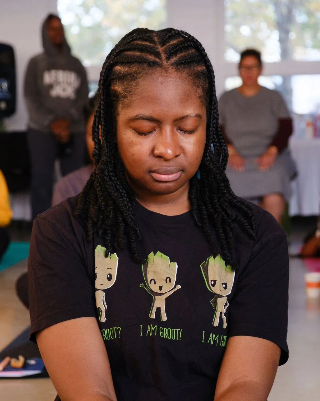 A young woman with braided hair sitting with her eyes closed, wearing a black T-shirt with cartoon characters of Groot from Guardians of the Galaxy and the text 'I am Groot!'