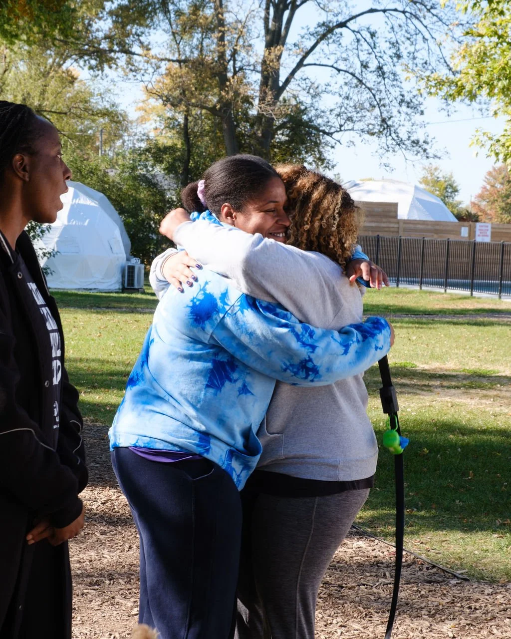 Two women hugging each other tightly and smiling, outside on a sunny day, with trees and structures in the background. One woman has her eyes closed and is embracing the other, who has curly hair and a walking stick. Another woman is standing nearby,