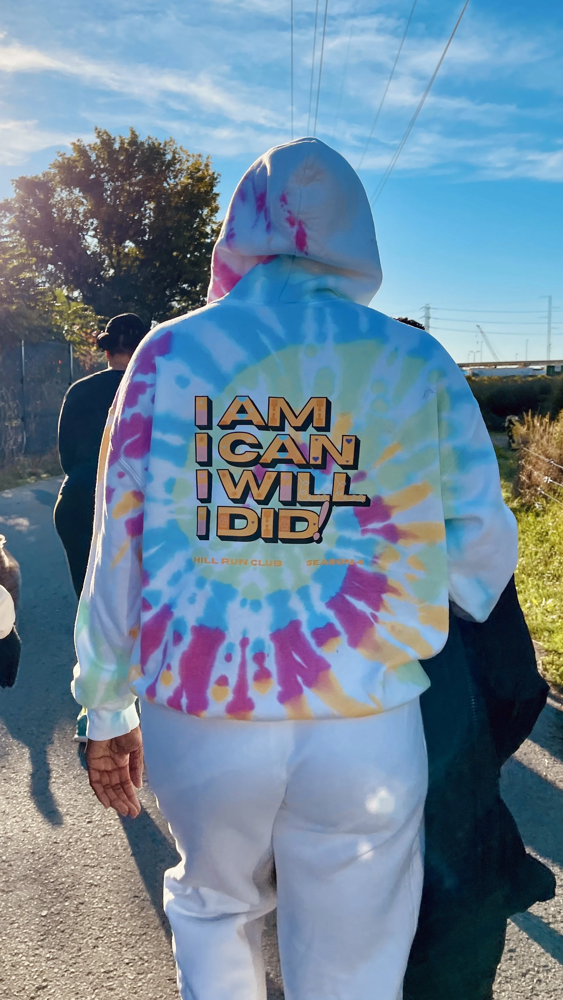 Person wearing a colorful tie-dye hoodie with motivational text walking outdoors during daytime.