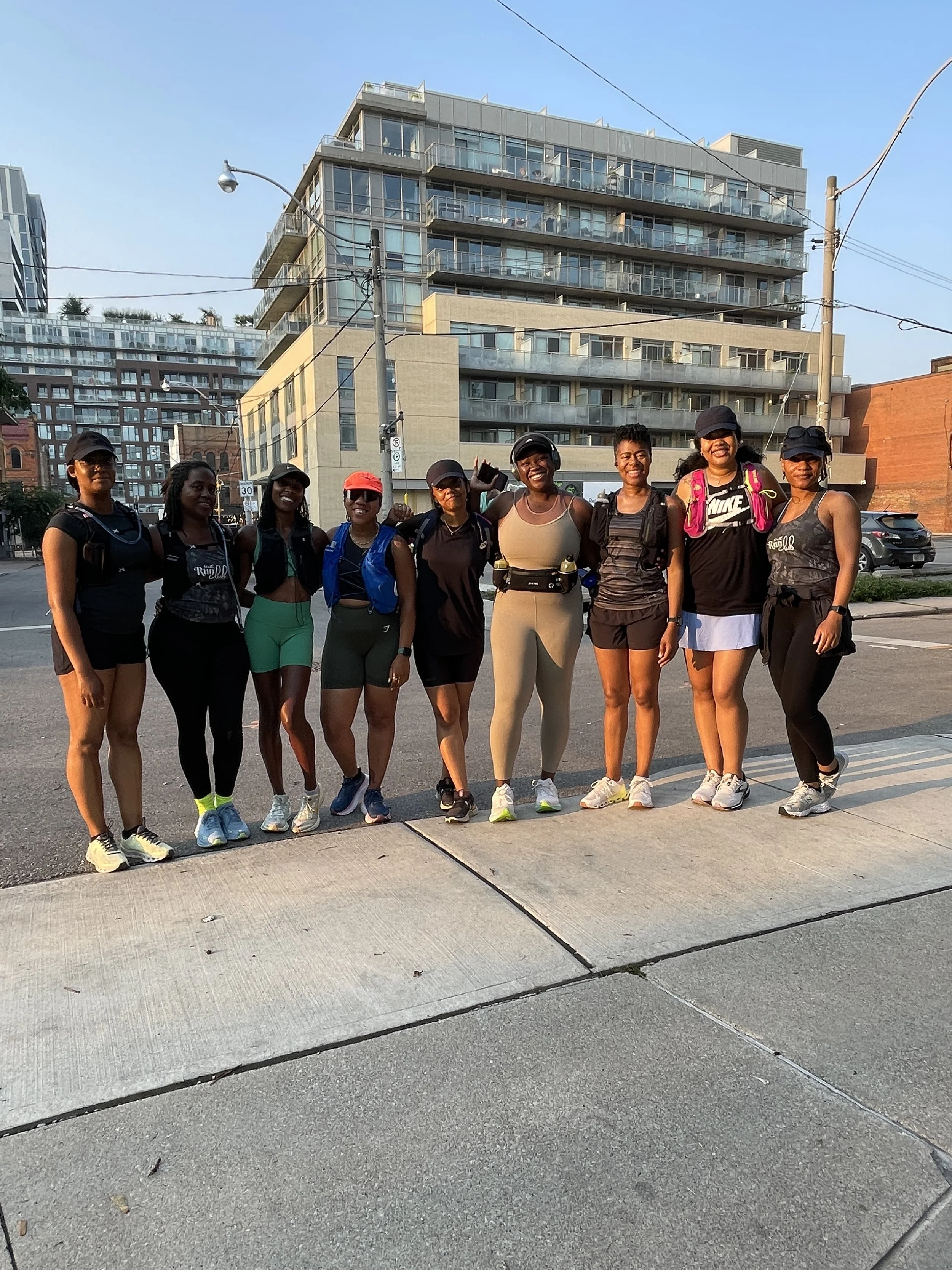 Group of nine women in athletic wear standing on sidewalk in urban area, smiling, with modern apartment buildings in the background.