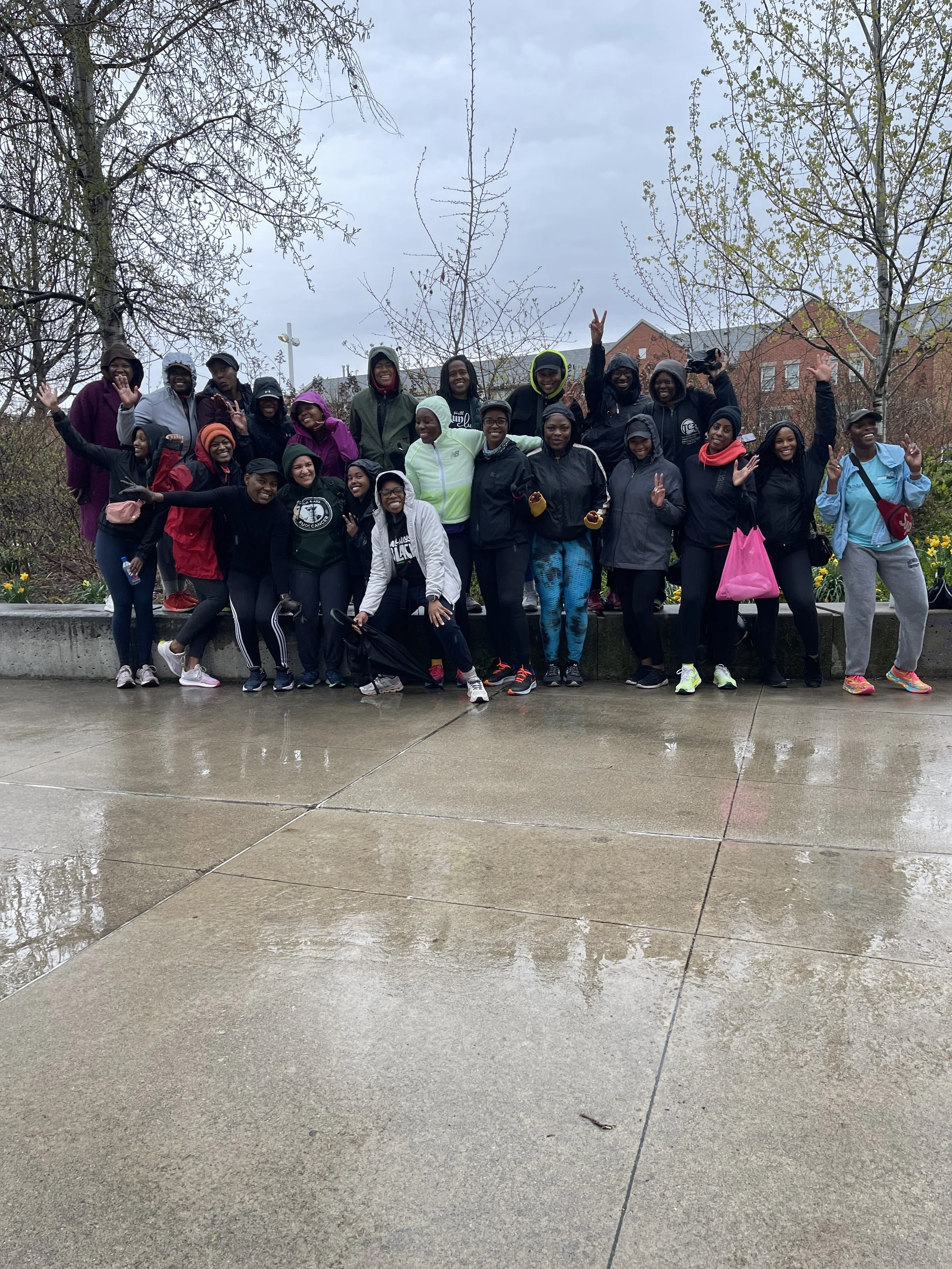 A large group of people outdoors in rainy weather, smiling and posing for a group photo on a wet pavement with wet trees and overcast sky in the background.