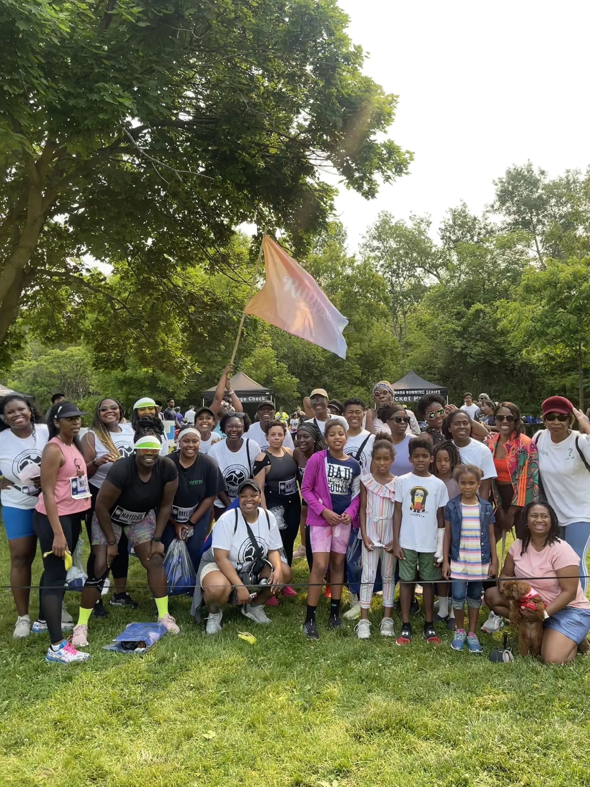 A group of diverse people gathered outdoors in a park with trees, posing for a photo. Some individuals are smiling, wearing athletic clothing, and holding a flag. There are children, adults, and a person with a small dog, indicating a community or fa