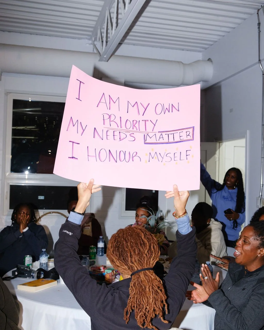 A woman with long reddish-brown dreadlocks holds a pink poster board with a handwritten message at a gathering. The message reads, 'I AM MY OWN PRIORITY MY NEEDS MATTER I HONOR MYSELF,' with colorful letters and stars. She is surrounded by seated wom