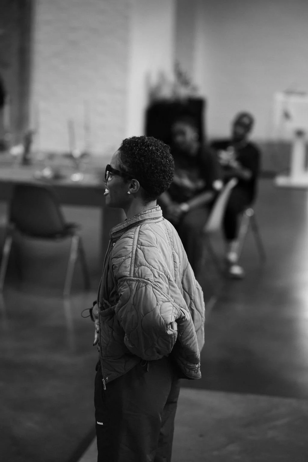 A woman with short curly hair and glasses smiling, wearing a quilted jacket, standing in focus. In the background, two people are seated at a table, slightly out of focus.