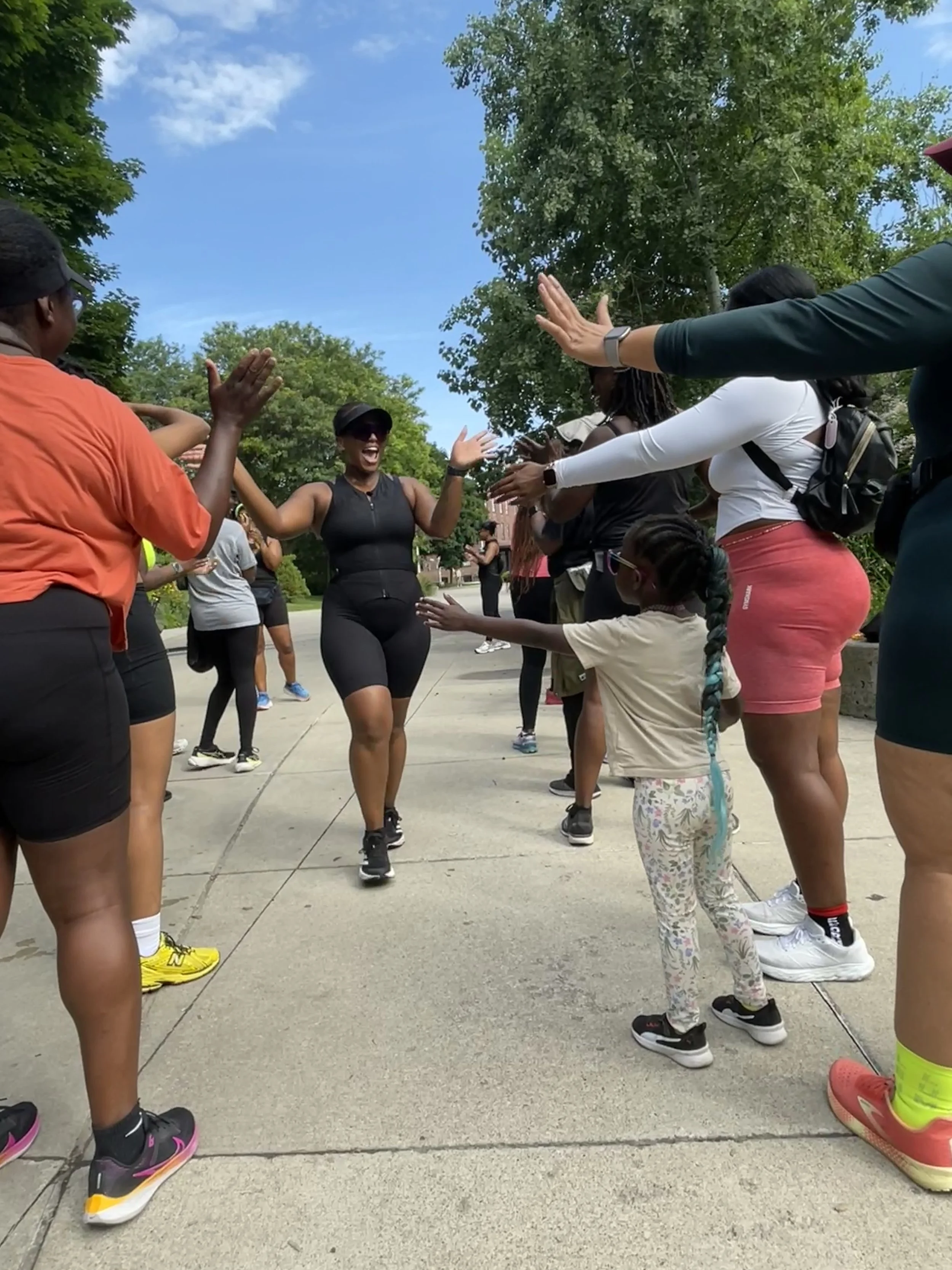 A group of people outdoors, forming a circle and clapping, with a woman in the center smiling and running through the circle. The scene takes place on a sunny day with trees and a blue sky in the background.