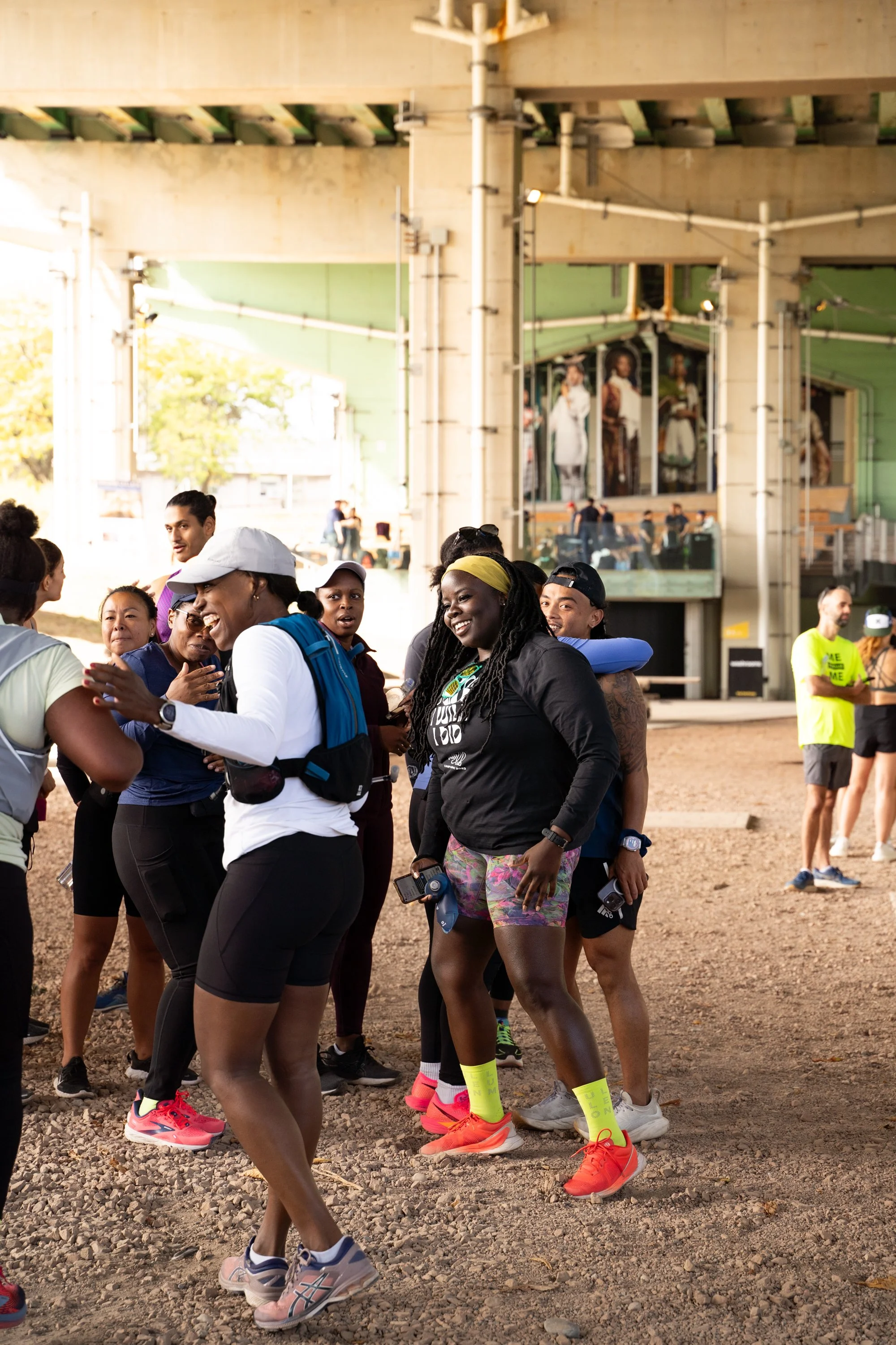 Group of people gathered outdoors under a bridge, smiling and talking, with some wearing athletic clothing and running shoes. The background shows an underpass with graffiti and some onlookers sitting on a balcony.
