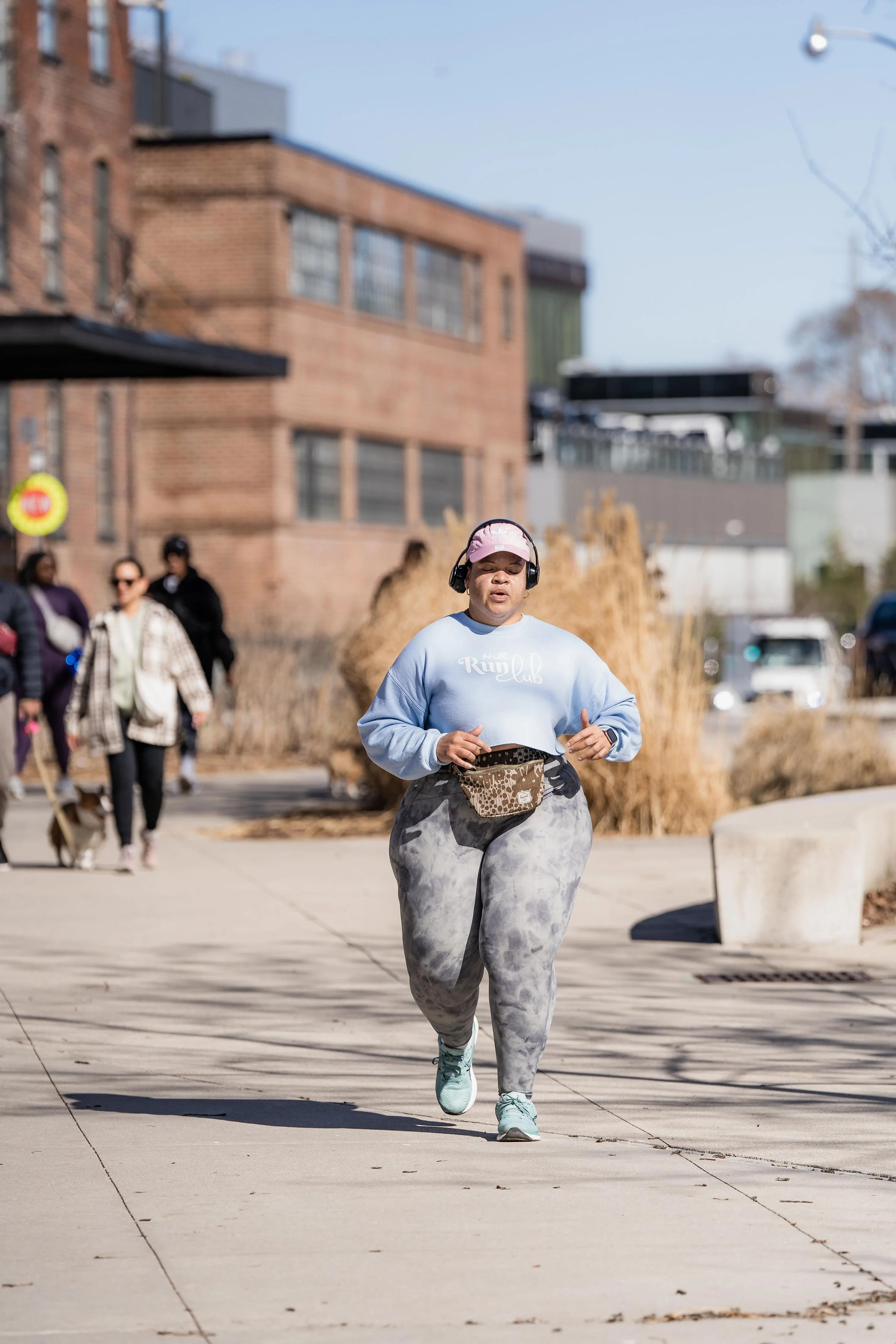 A woman jogging on a city sidewalk with other pedestrians in the background on a sunny day.
