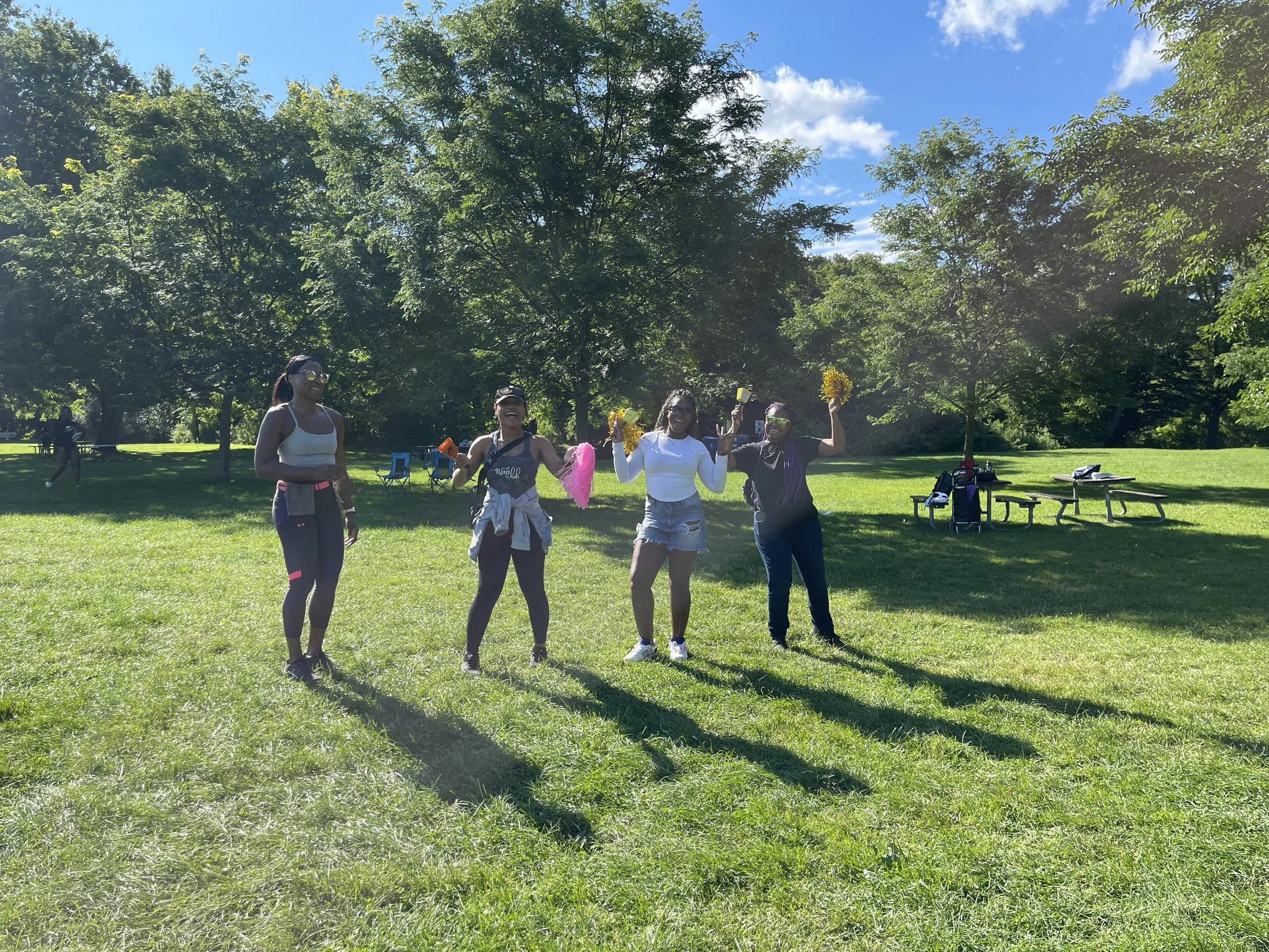 Four women standing on a grassy field in a park with trees in the background, enjoying a sunny day with some holding yellow pom-poms and pink pom-poms.