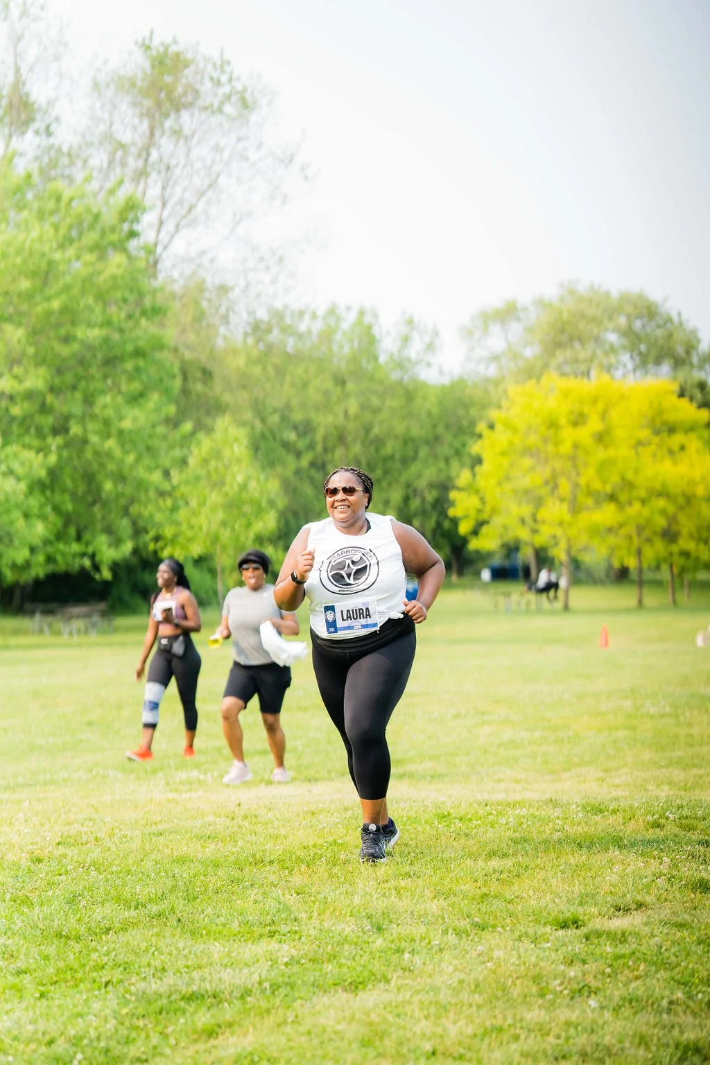 A woman running outdoors during a race, wearing sunglasses, a white tank top with a logo, black leggings, and a bib with the name 'Laura.' She is smiling and appears to be enjoying the event, with other women running behind her on a grassy field with