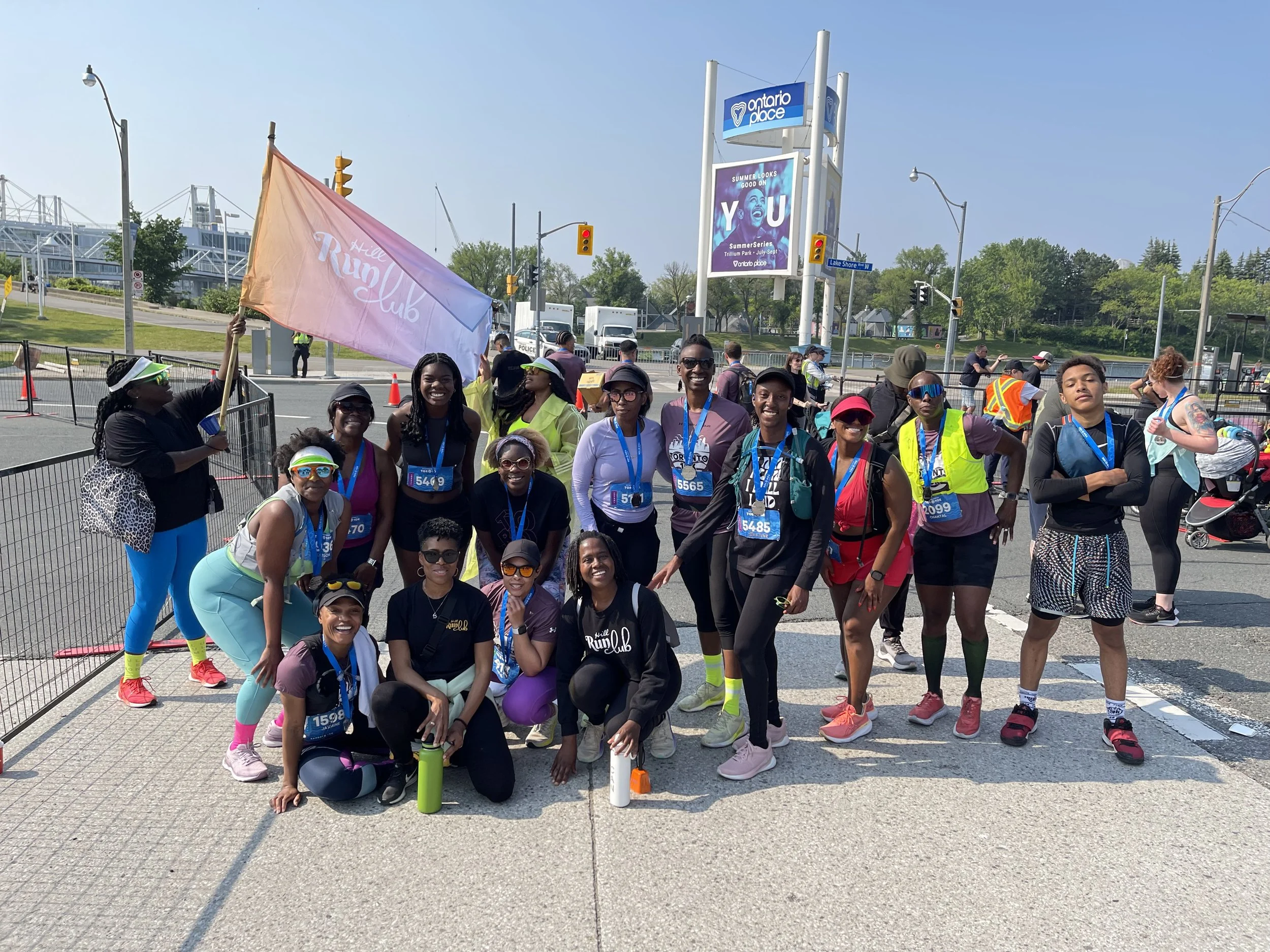 A group of diverse runners wearing medals and race bibs pose for a photo after completing a race on a city street, with some holding a pink flag that says 'This Run Club.'