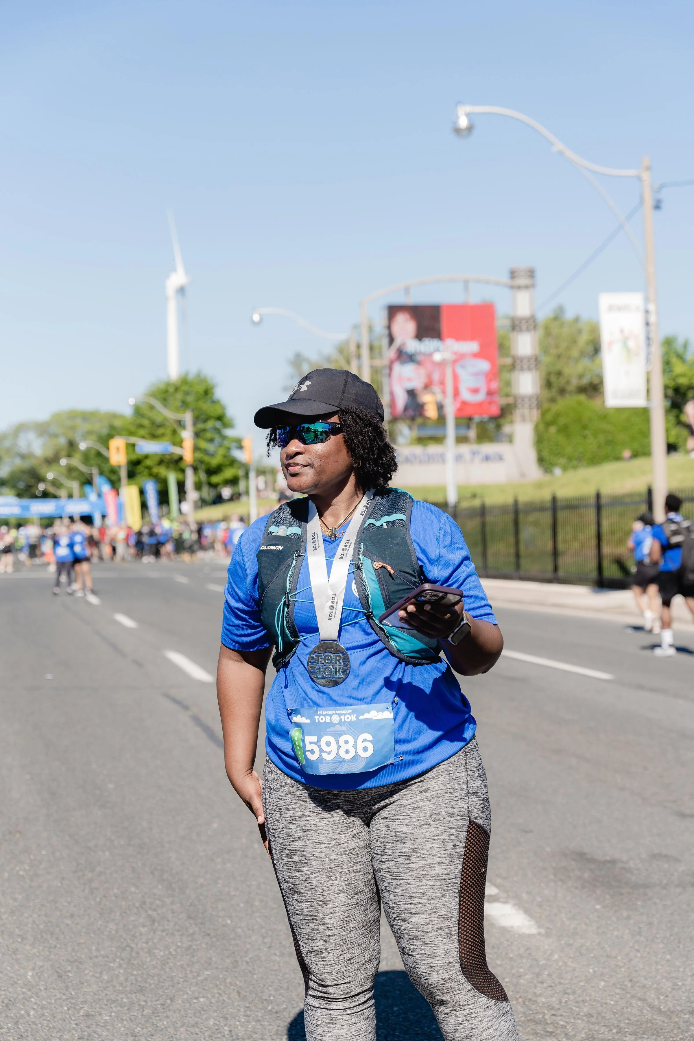 A woman at a marathon event, wearing a blue shirt, gray leggings with black mesh panels, sunglasses, a black cap, and a backpack, holding a phone, with a medal hanging around her neck.