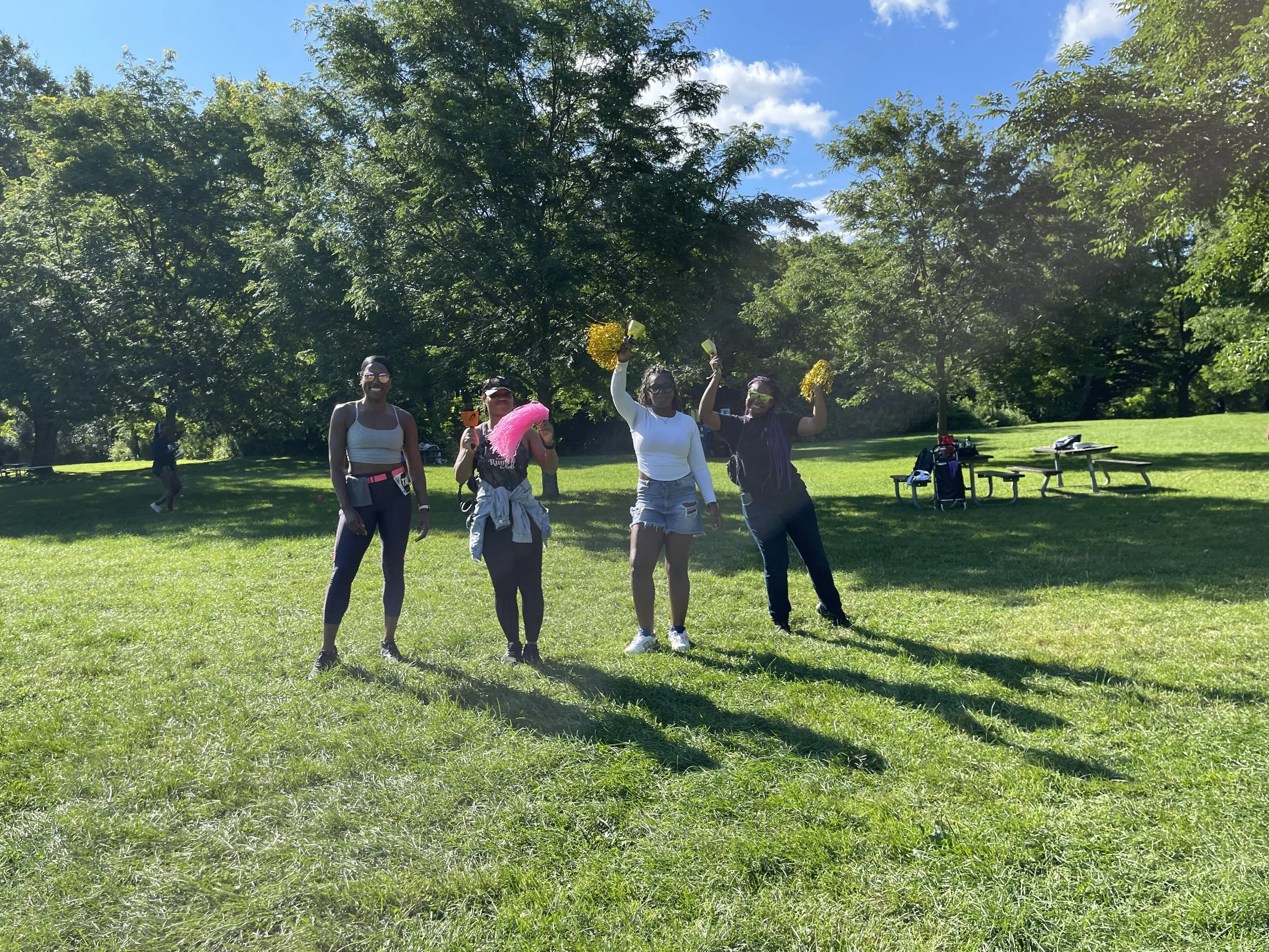 Four women standing on a grassy field in a park, smiling and celebrating with the sun shining and trees in the background.