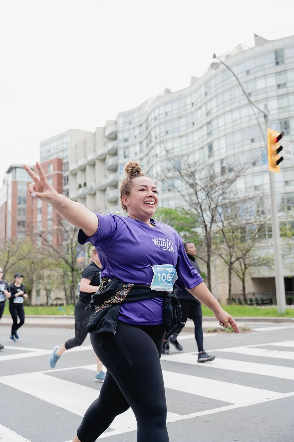 A woman running in a race, smiling and making a peace sign with her hand, wearing a purple shirt that says 'Run Club', with other runners in the background in an urban setting with buildings and a traffic light.